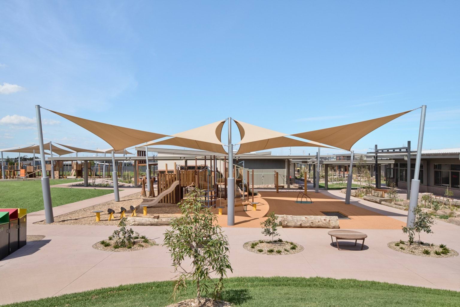 An outdoor play space with climbing equipment, shade sails, curved paths, and newly planted garden beds.