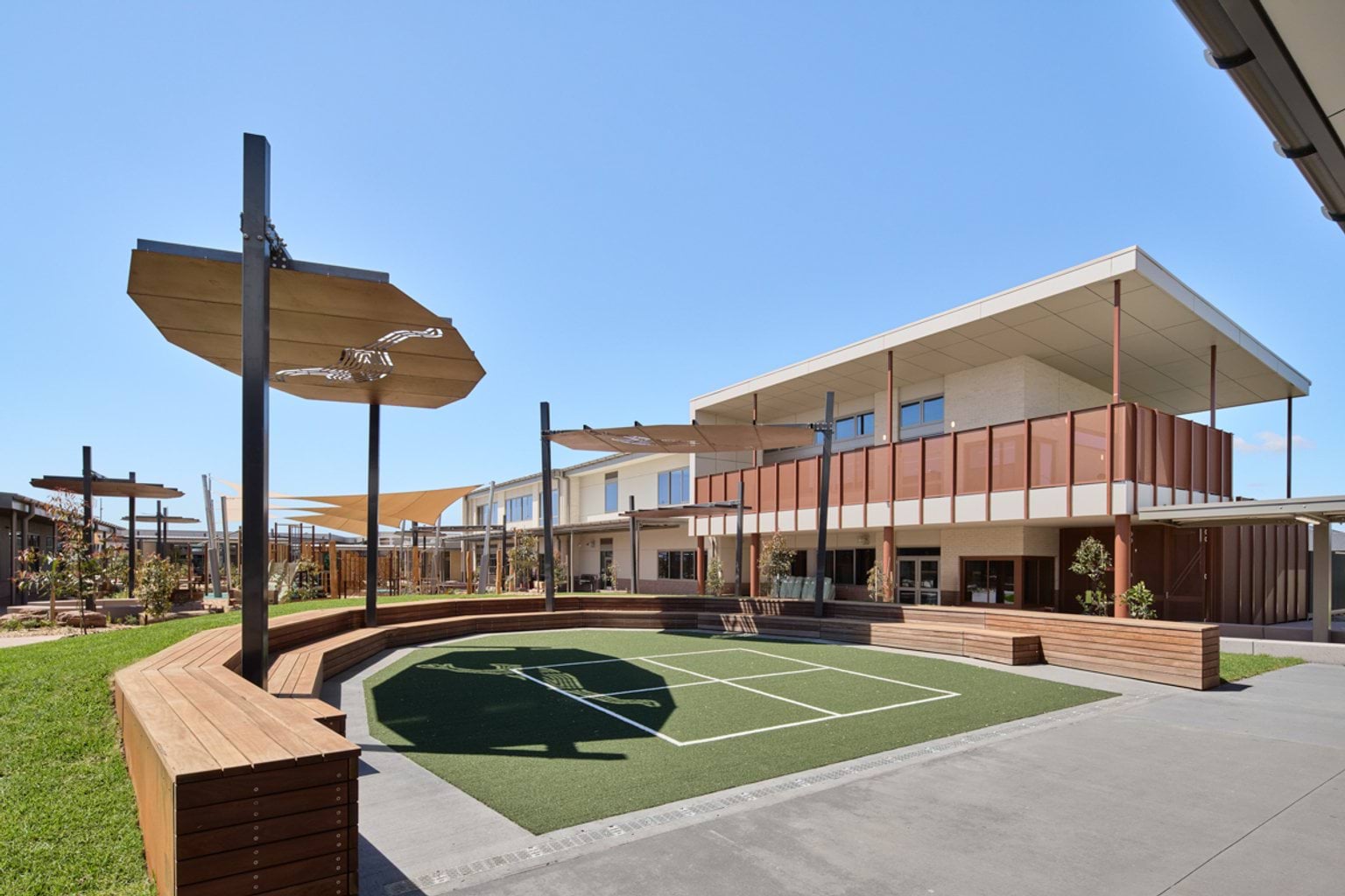 An outdoor courtyard with tiered timber seating, shade structures, and a two‑storey school building in the background.