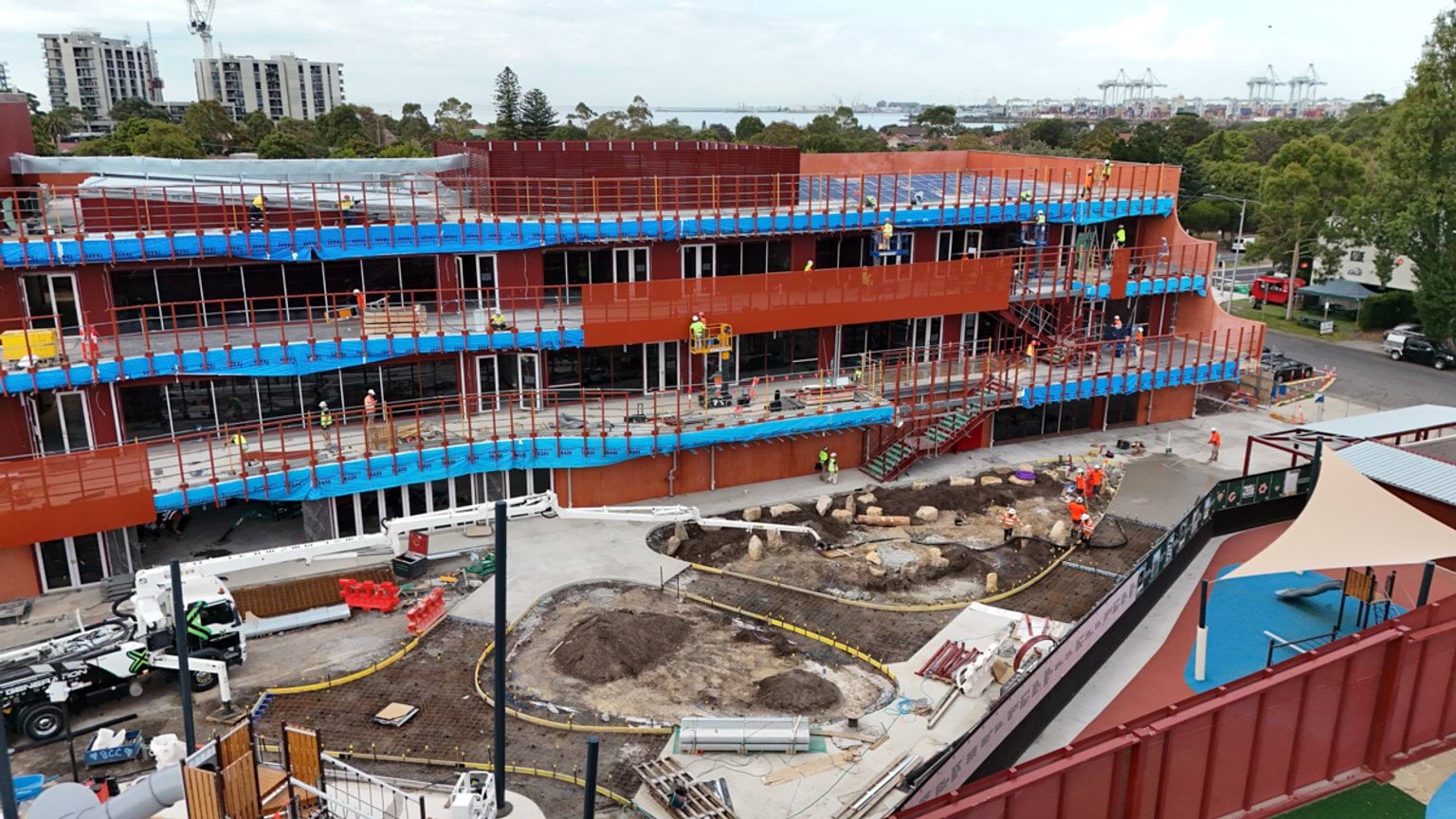 A construction site view of the school building showing red exterior panels, scaffolding, and landscaping works underway.