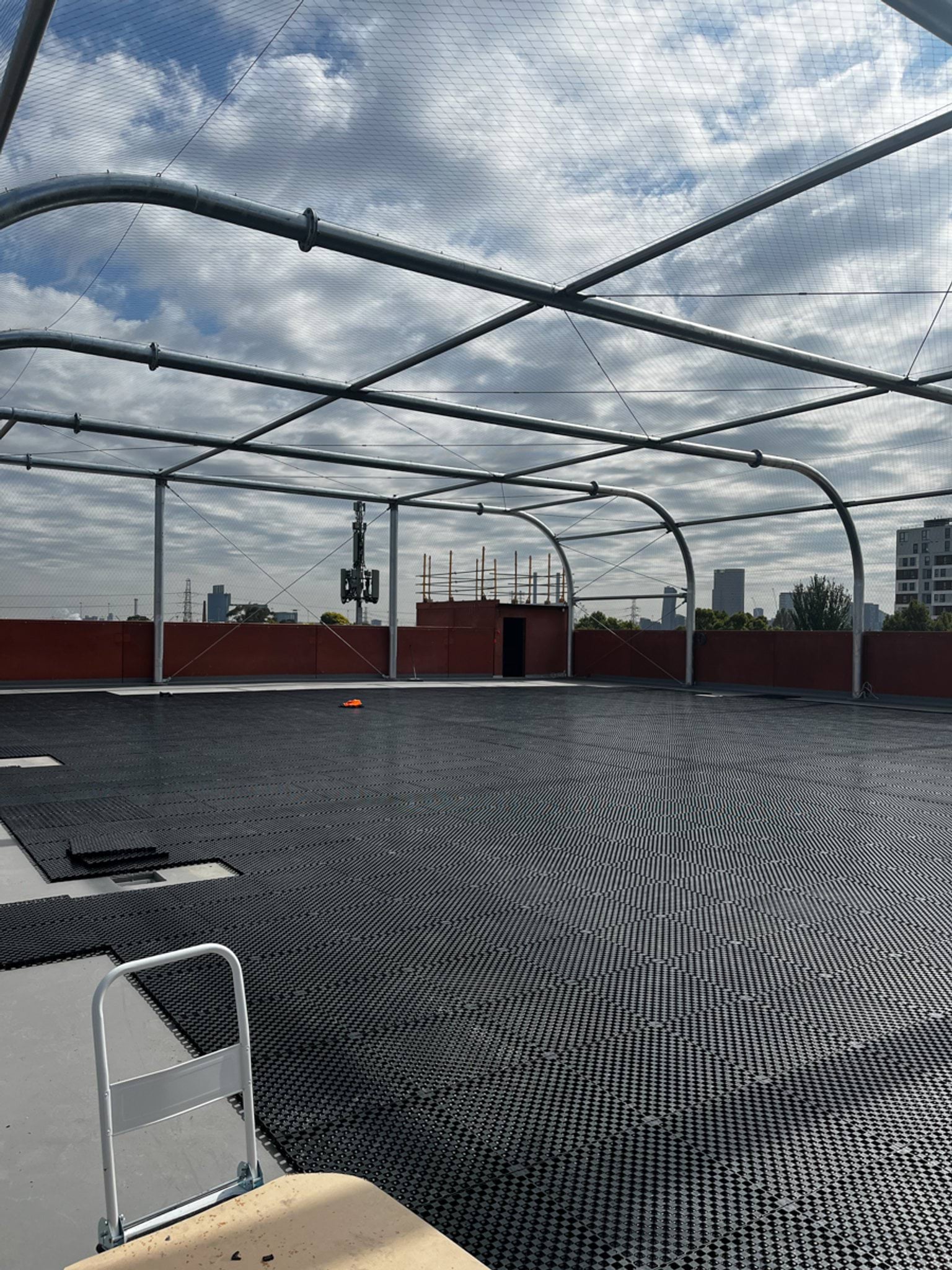 A rooftop play space under construction with curved steel net supports and textured flooring tiles.