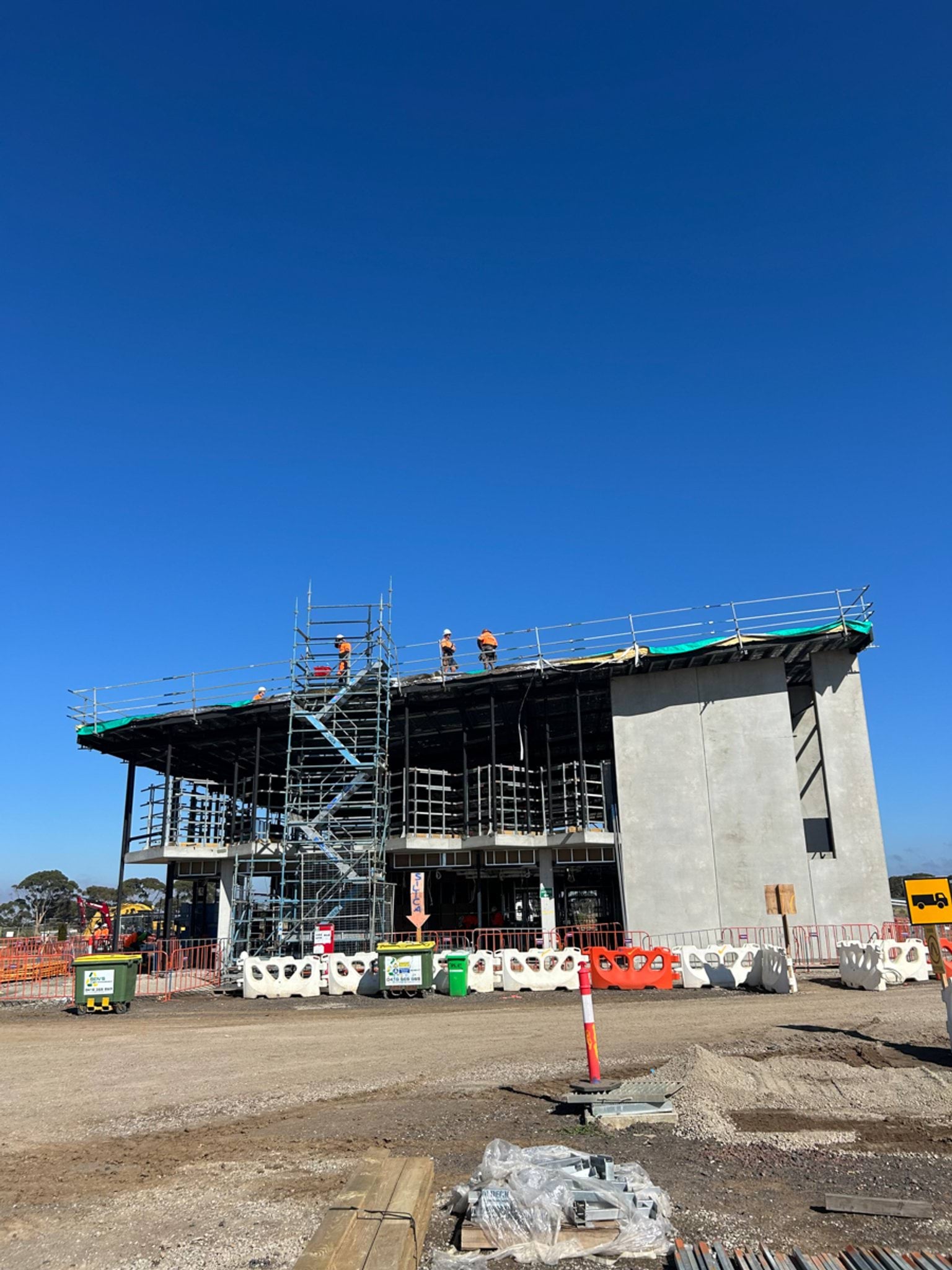 A partially constructed two‑storey school building with scaffolding, workers on the roof, and construction barriers in the foreground.