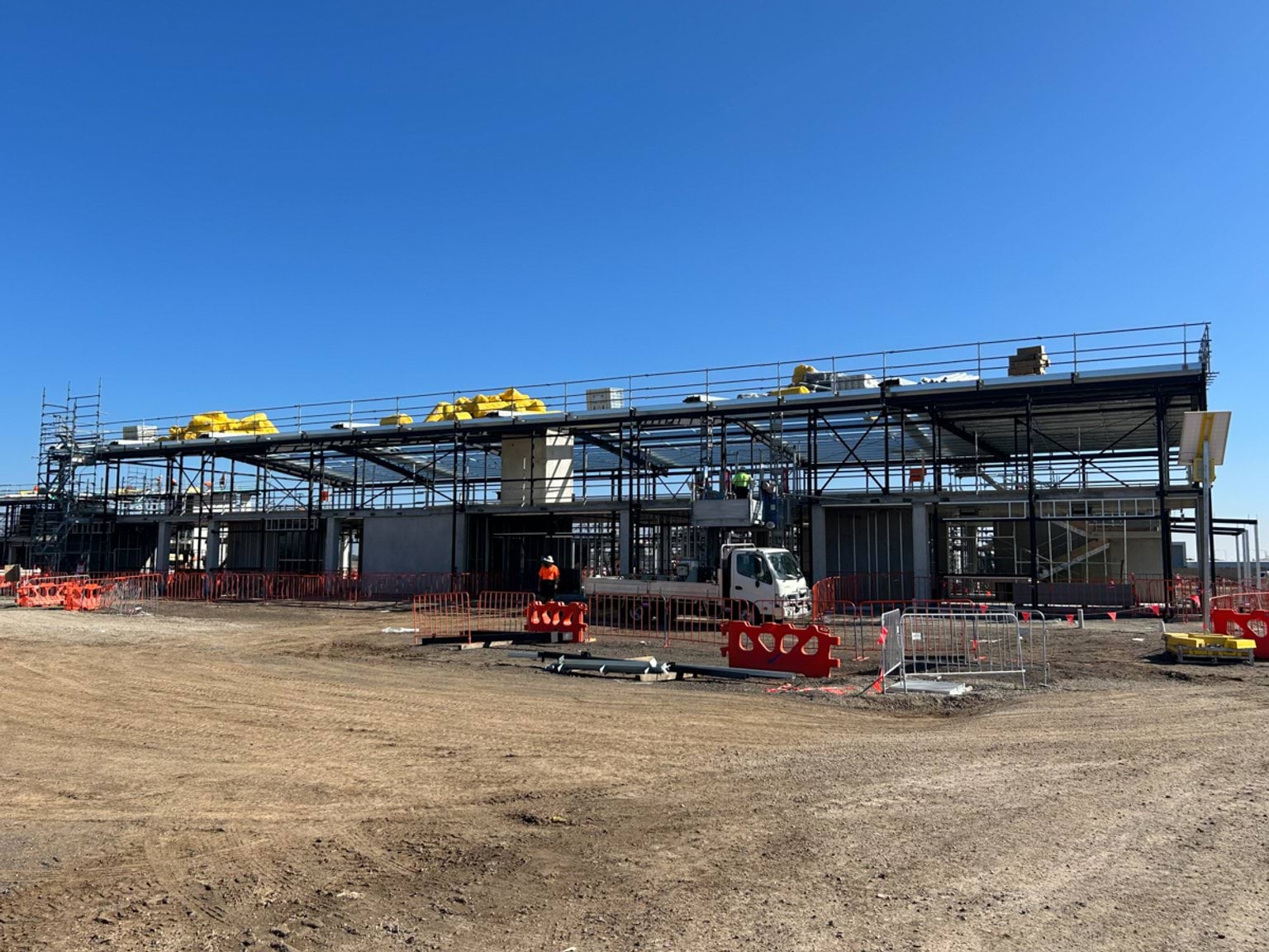 A long steel‑framed school building under construction with cranes, scaffolding, and workers on site.