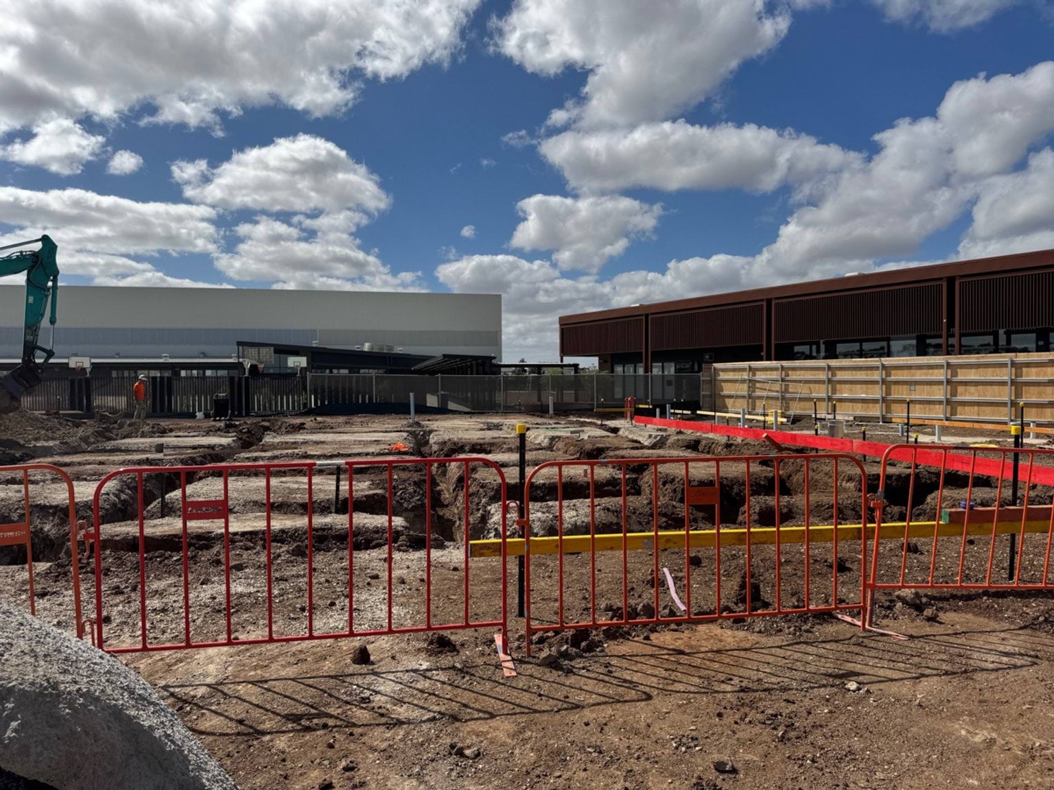 Excavated trenches and groundwork within a fenced construction area, with machinery and nearby school buildings in the background.