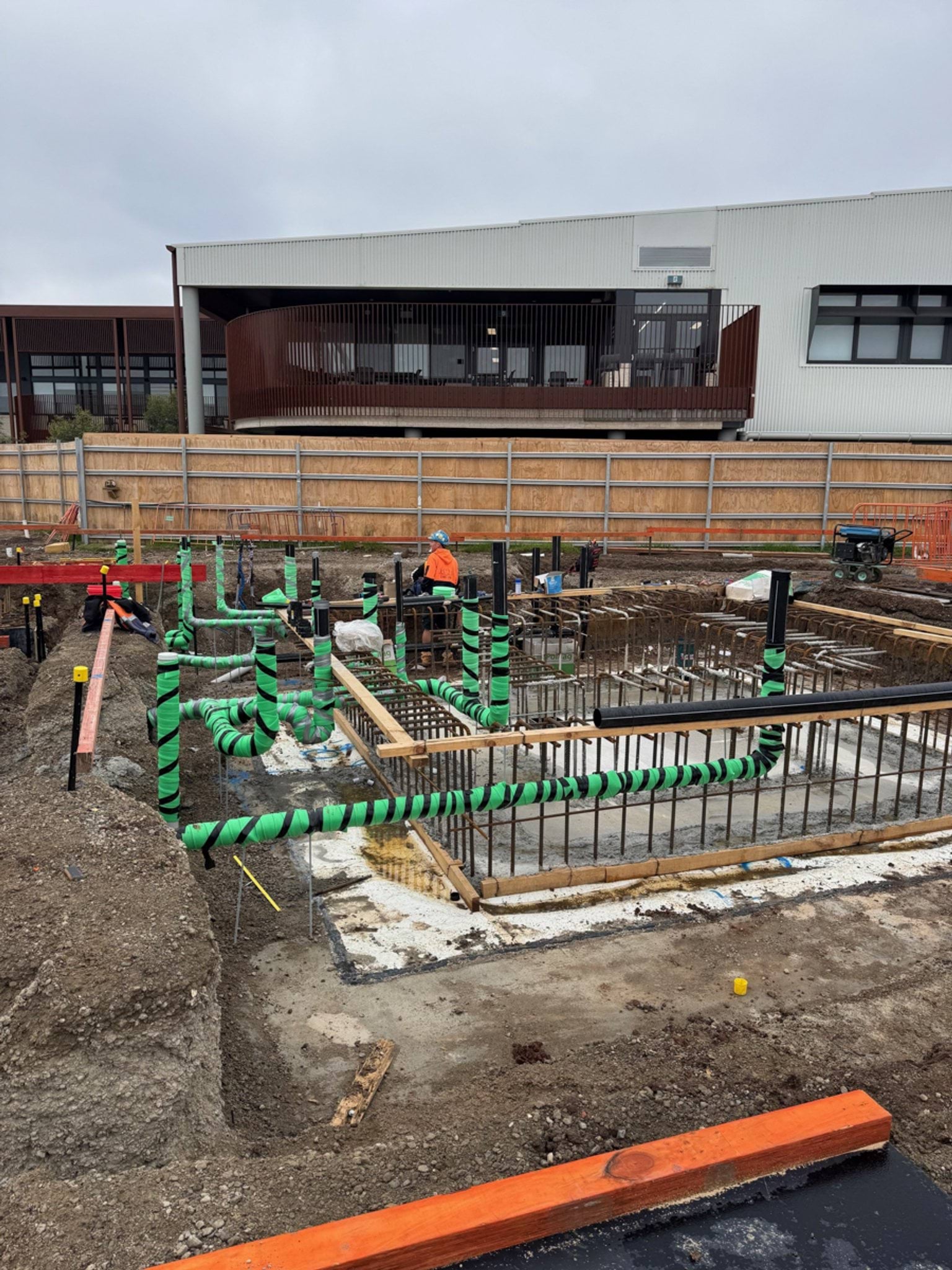 Workers installing services and reinforcement in an excavated foundation area, with a school building visible behind the site fencing.