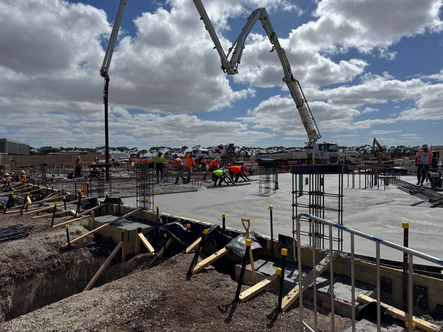 Concrete being poured onto a large foundation slab, with workers and pumping equipment active across the construction site.