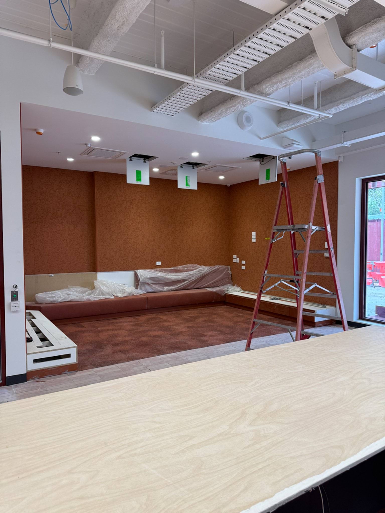 An indoor learning space under construction with built‑in seating along the back wall, plastic coverings, and a ladder in the centre of the room.