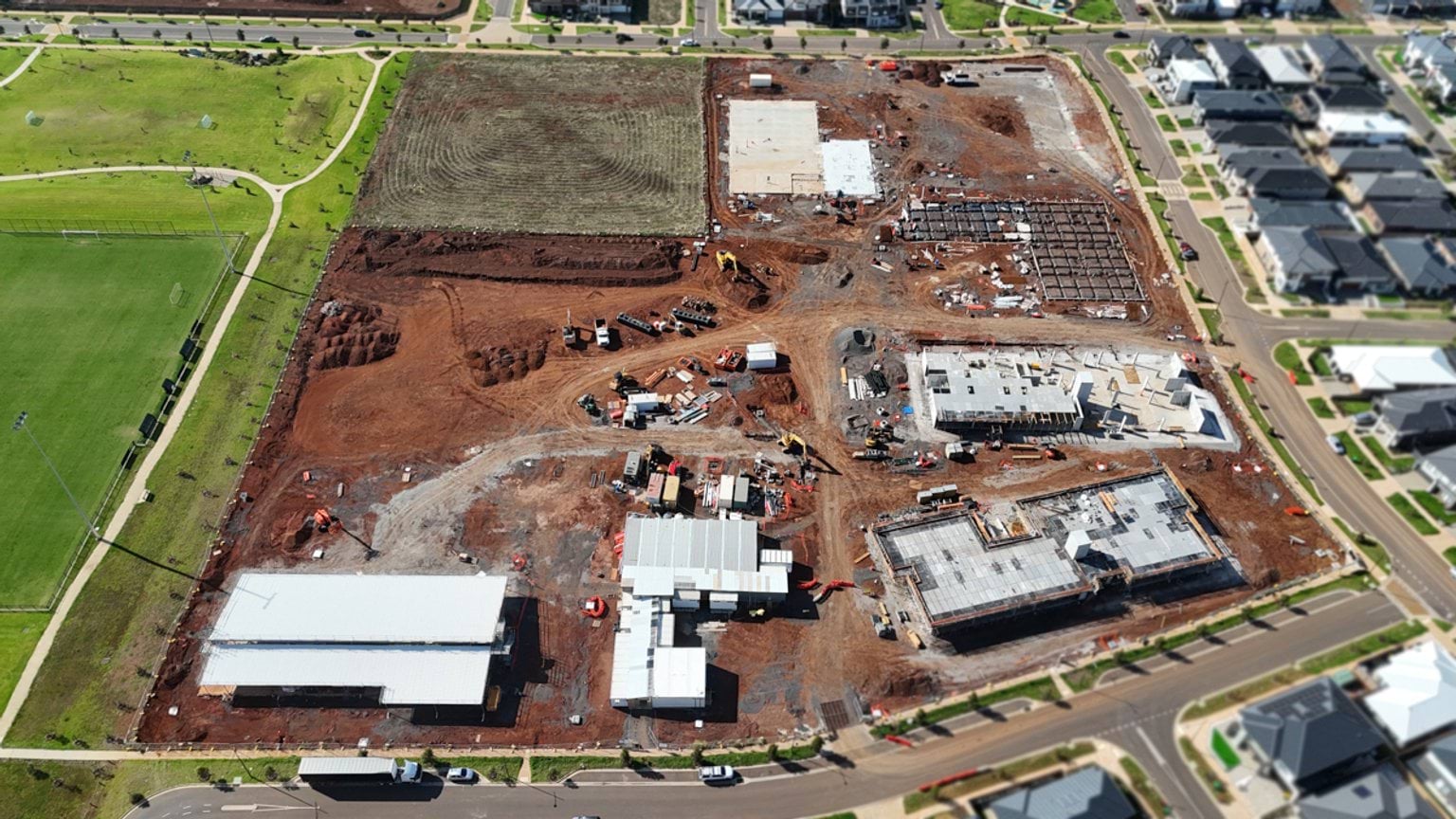 Wide aerial view of a kindergarten construction site beside sports fields, with multiple building footprints and earthworks underway.