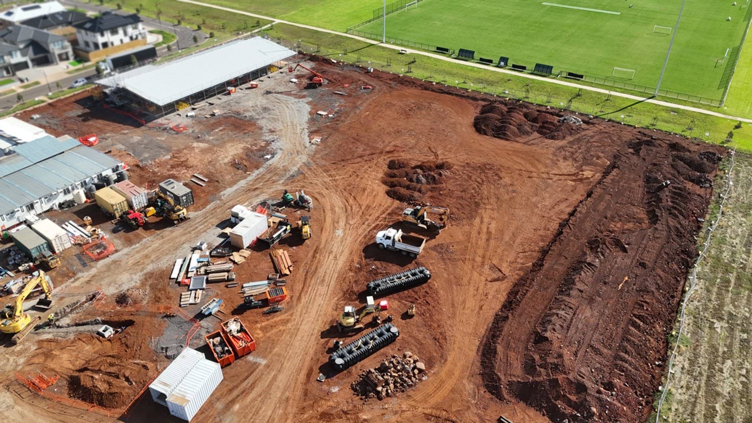 An aerial view of earthworks and material storage areas at the school site, with excavators and trucks operating on compacted soil.