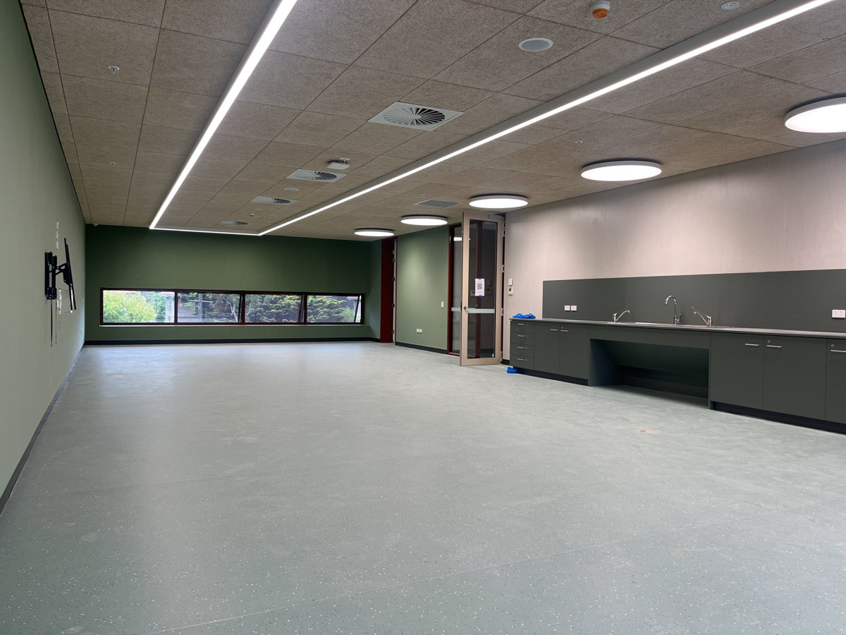 A large multi‑purpose learning space with green feature walls, vinyl flooring, ceiling lights, and a kitchenette along one wall.