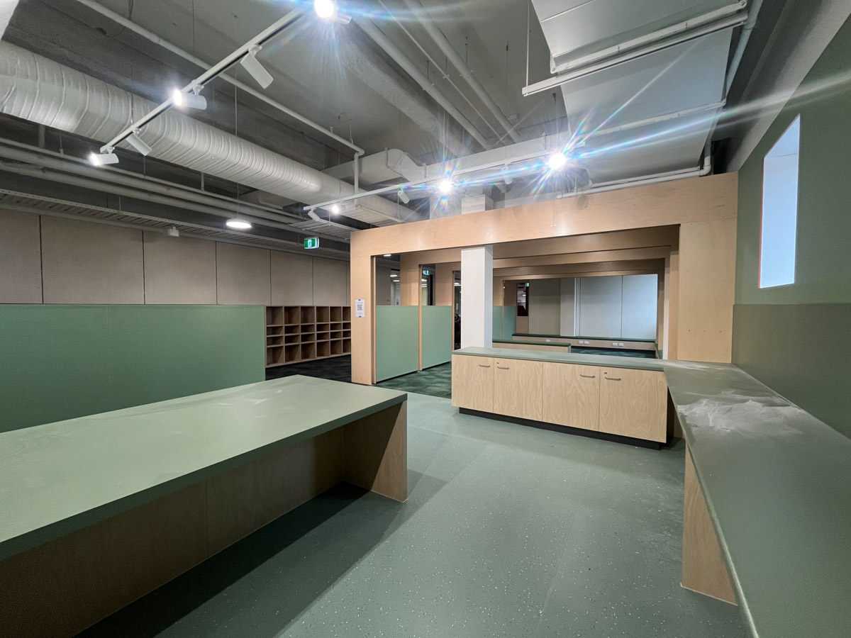 A practical learning space with timber benches, storage cupboards, green work surfaces, and exposed ceiling ductwork.