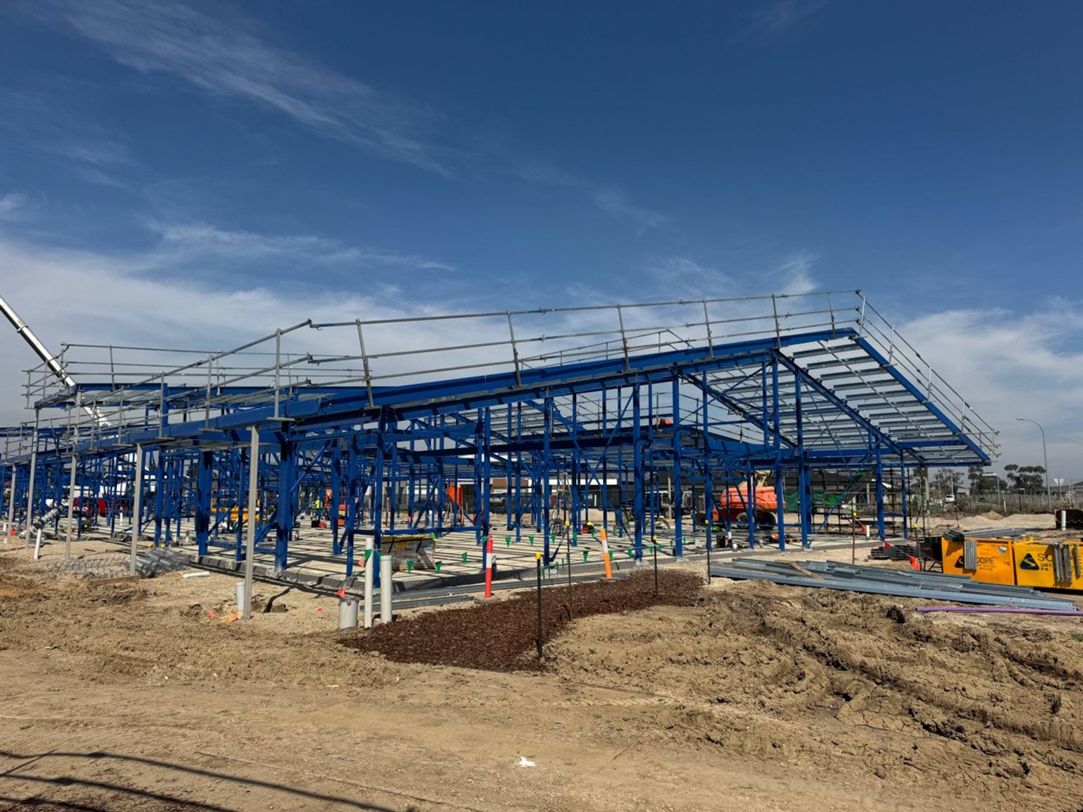 A large steel structure forming part of a school building, with roof framing in place and foundation works visible below.