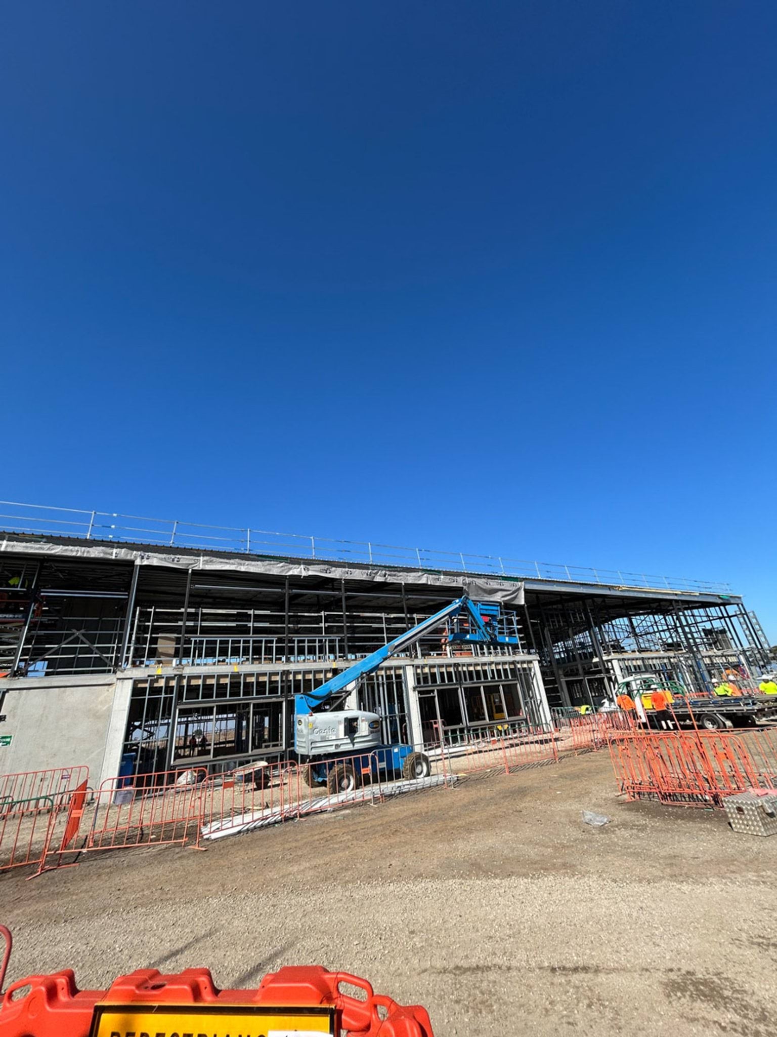 A partially enclosed school building under construction with steel framing, elevated work platforms, and workers operating on site.