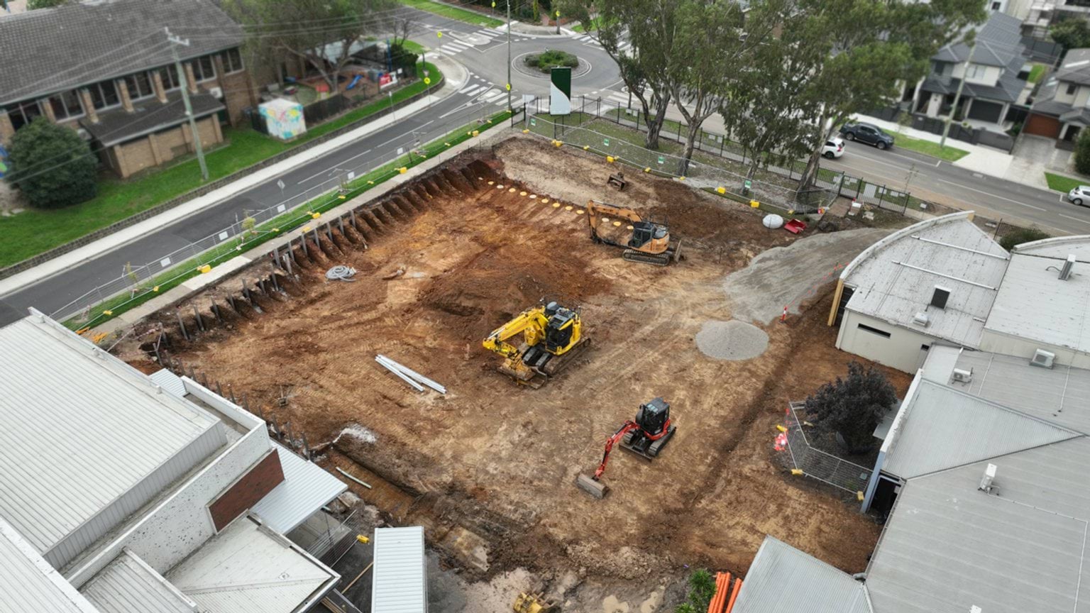 Overhead view of a fenced school construction area showing multiple excavators working within a large excavation.