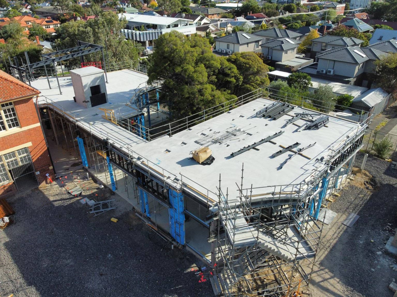 Aerial view of a school building under construction with a newly poured concrete roof slab, scaffolding and surrounding residential streets.