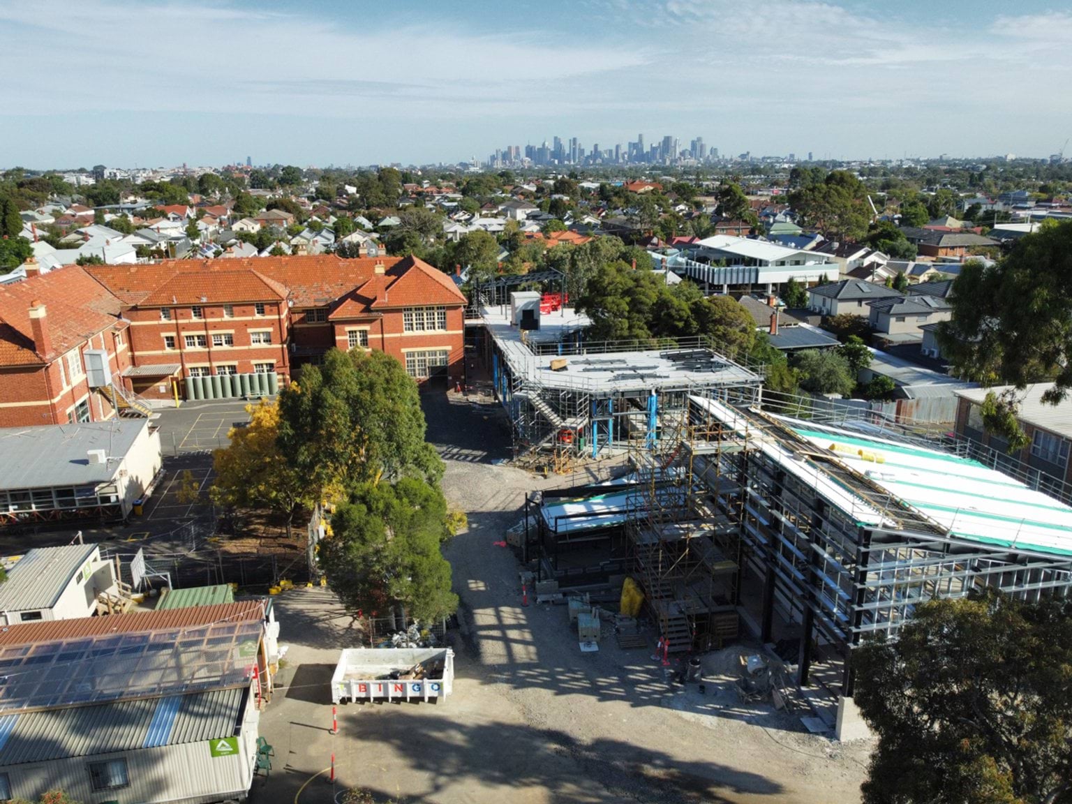 Wide aerial view of a school campus showing new building works alongside existing brick buildings, with the city skyline in the distance.