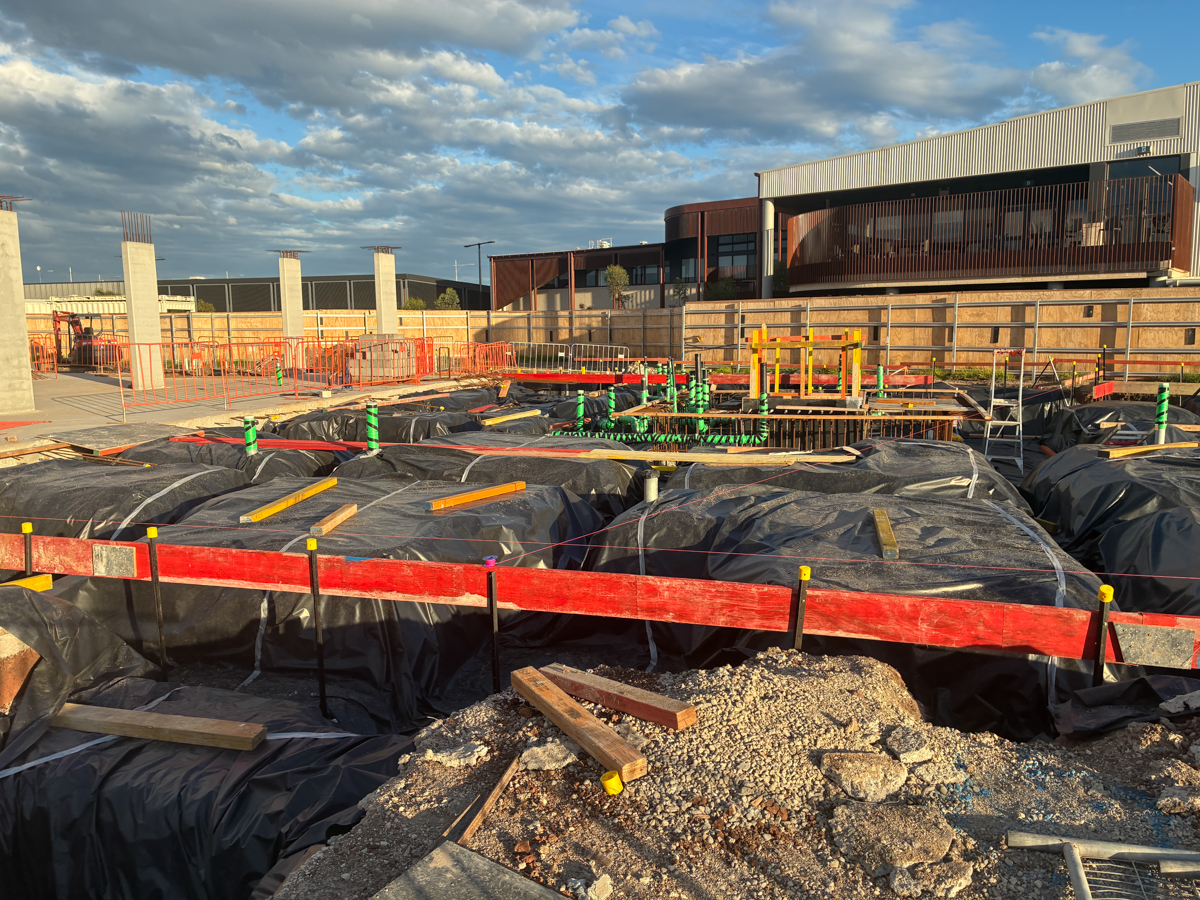 School building foundations with formwork, waterproofing and services installed, surrounded by safety fencing.