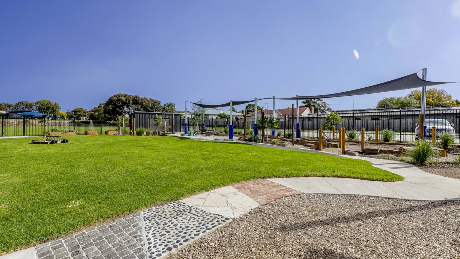 Open school playground with grassed area, shade sails, garden beds and nature‑based play elements.