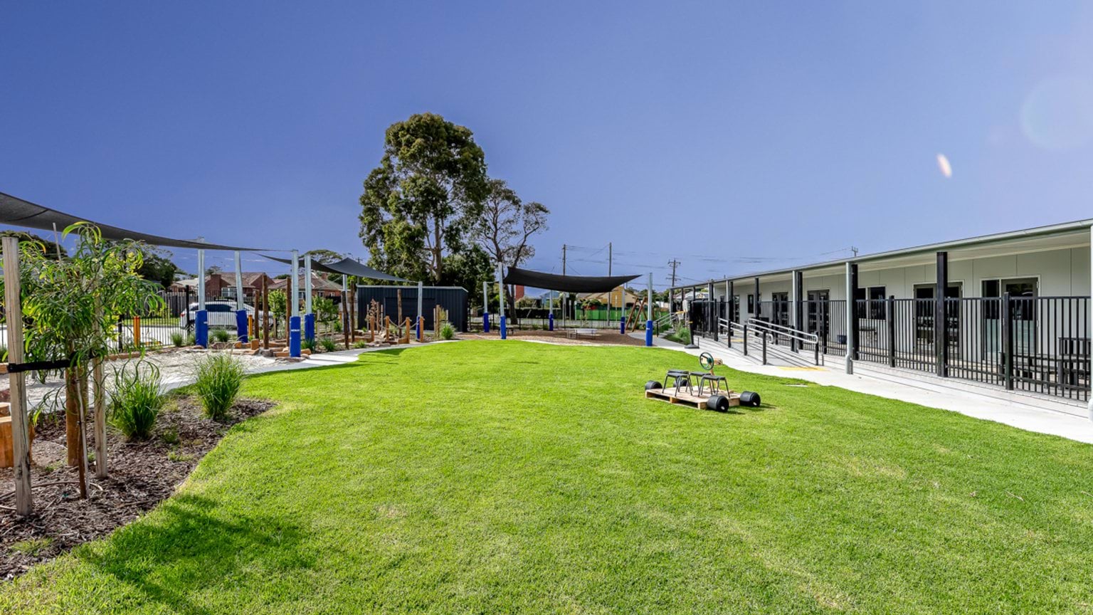 Wide view of a green school play space with ride‑on equipment, trees, shaded play structures and classroom buildings.