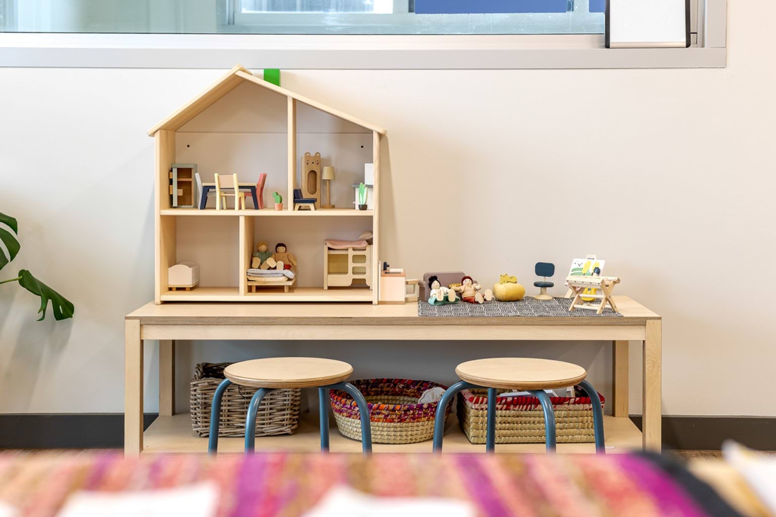 Wooden dollhouse play setup with figures and furniture on a low table with stools in an early learning room.