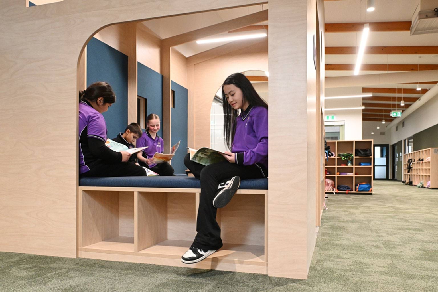 4 kids in purple and black school uniforms are sitting on bench seats inside a hallway area of their school. They're reading books and shelves with school bags are visible in the background.
