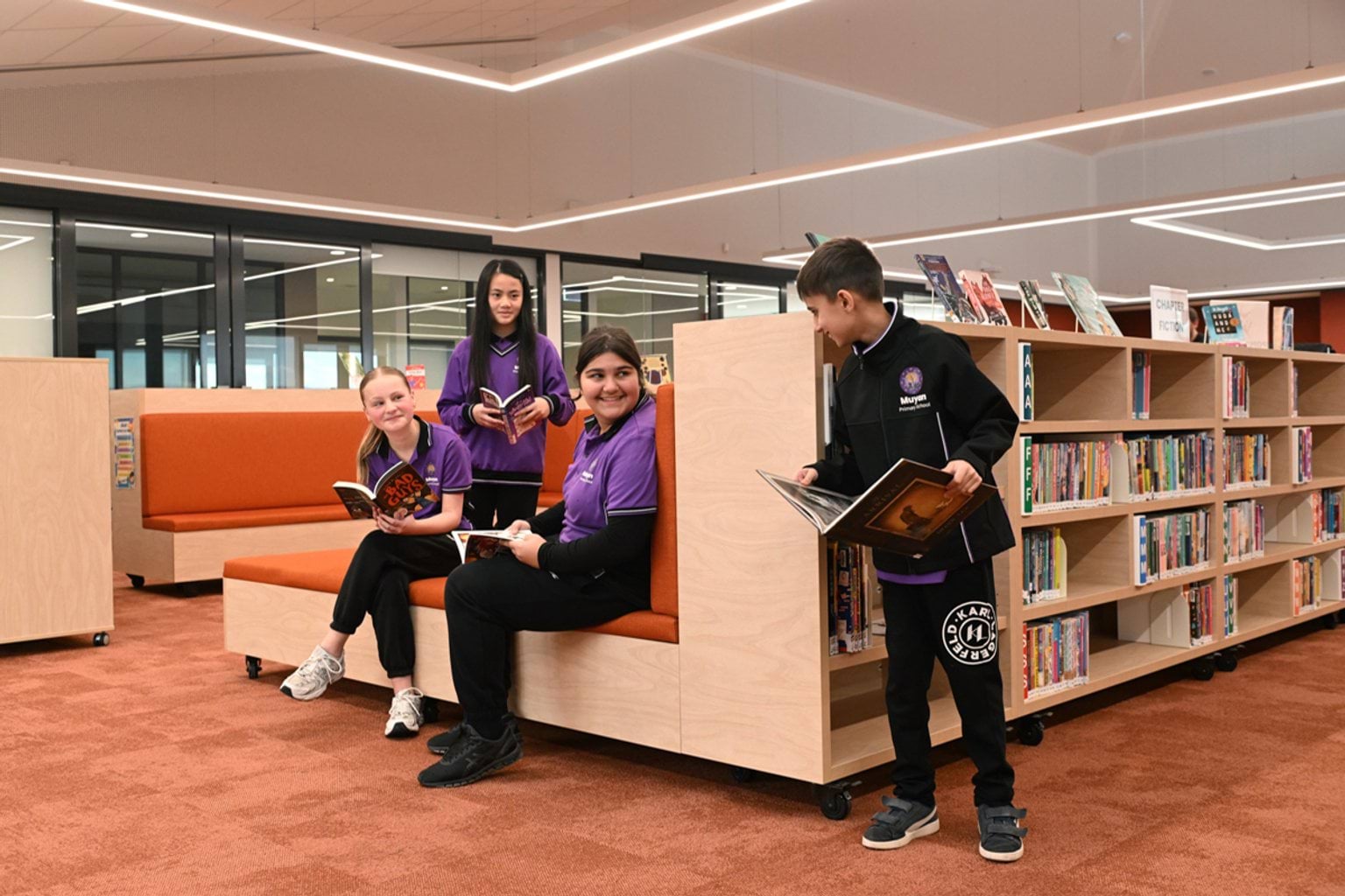 4 kids in purple and black school uniforms are inside a school library. 2 are seated holding books and 2 are standing holding books. There are shelves of books behind them.