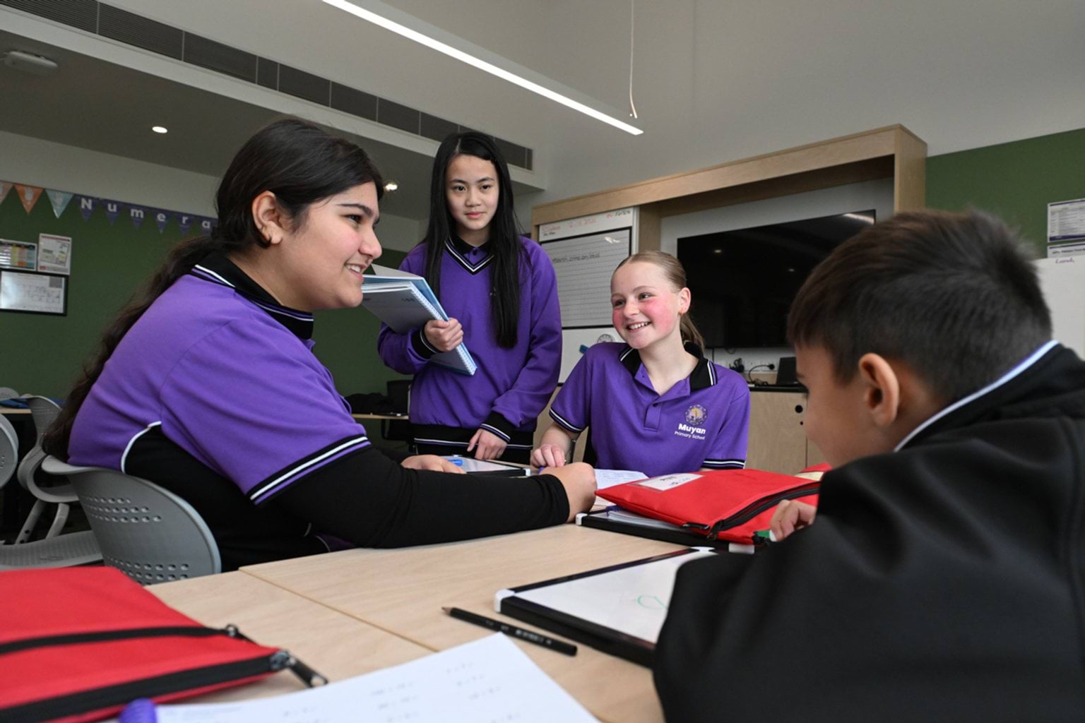 4 kids in purple and black school uniforms are gathered around desks in a classroom.