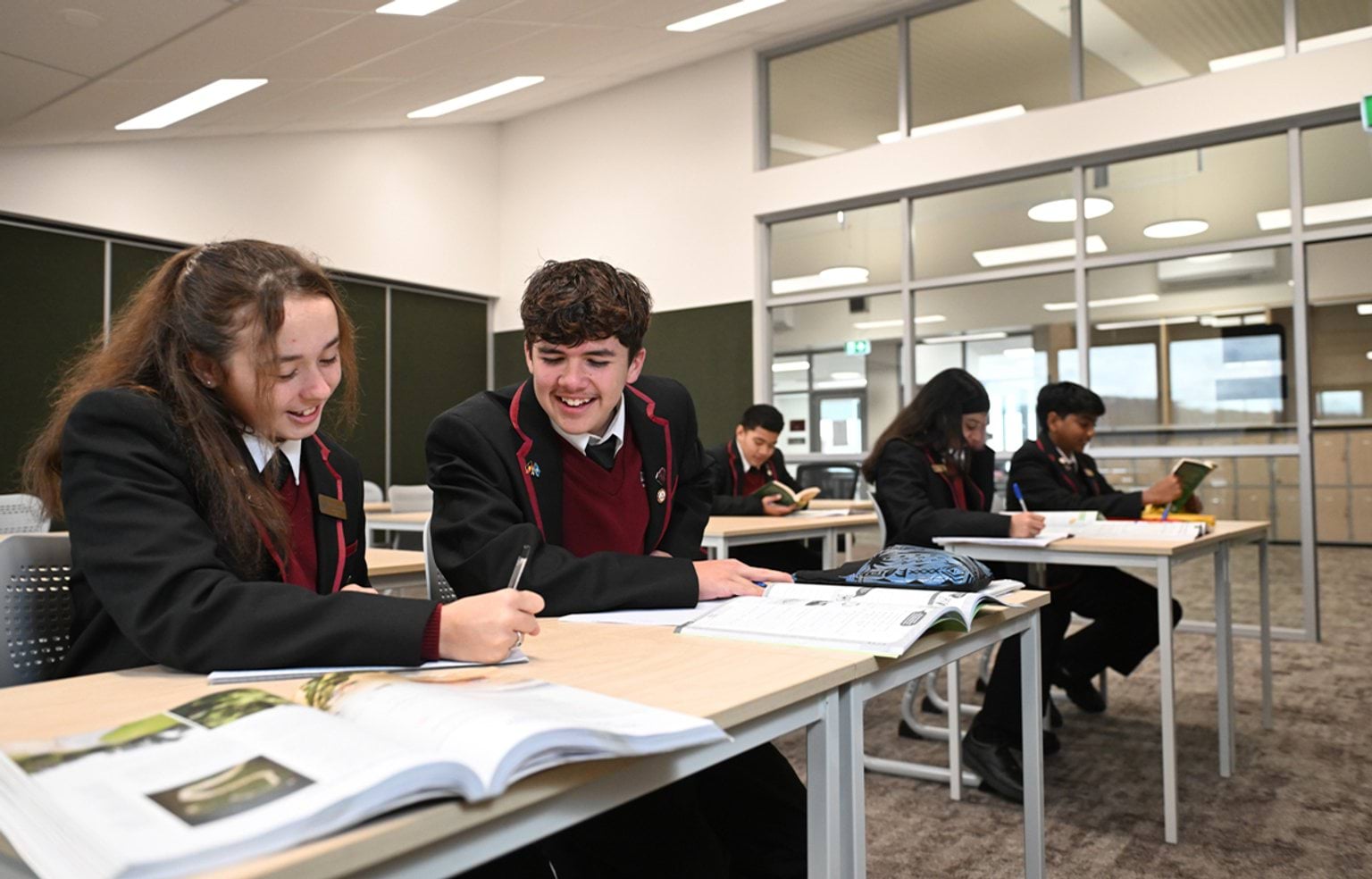 5 kids in maroon and black school uniform are seated in pairs at desks in a classroom, working in notebooks and reading textbooks. 