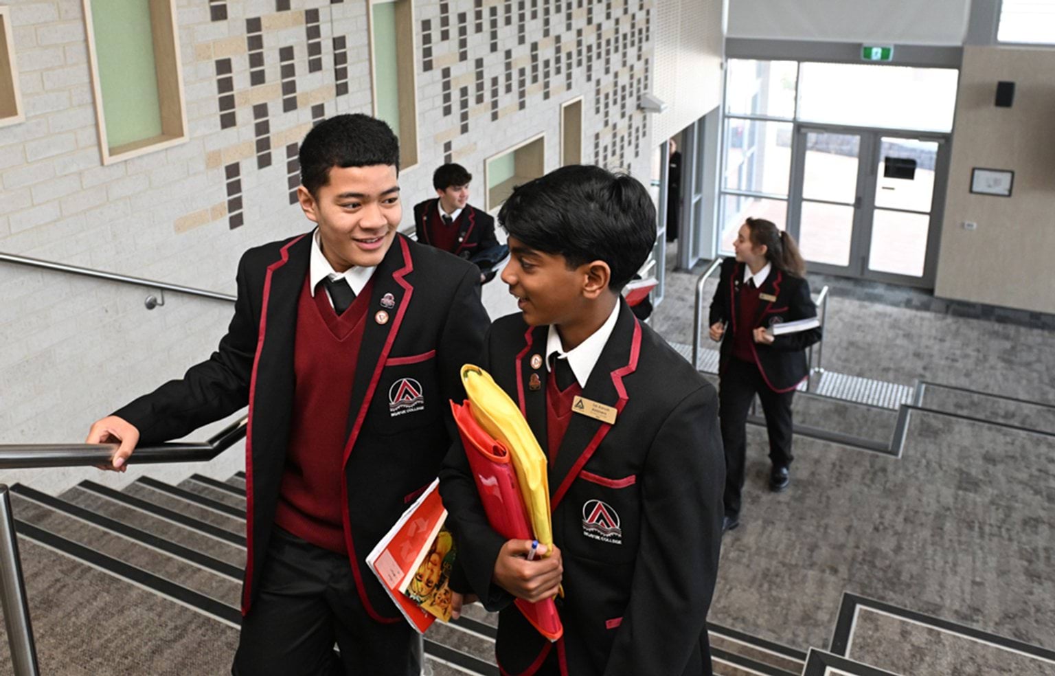 5 kids in maroon and black school uniform are walking up carpeted stairs inside their new school, holding school books.