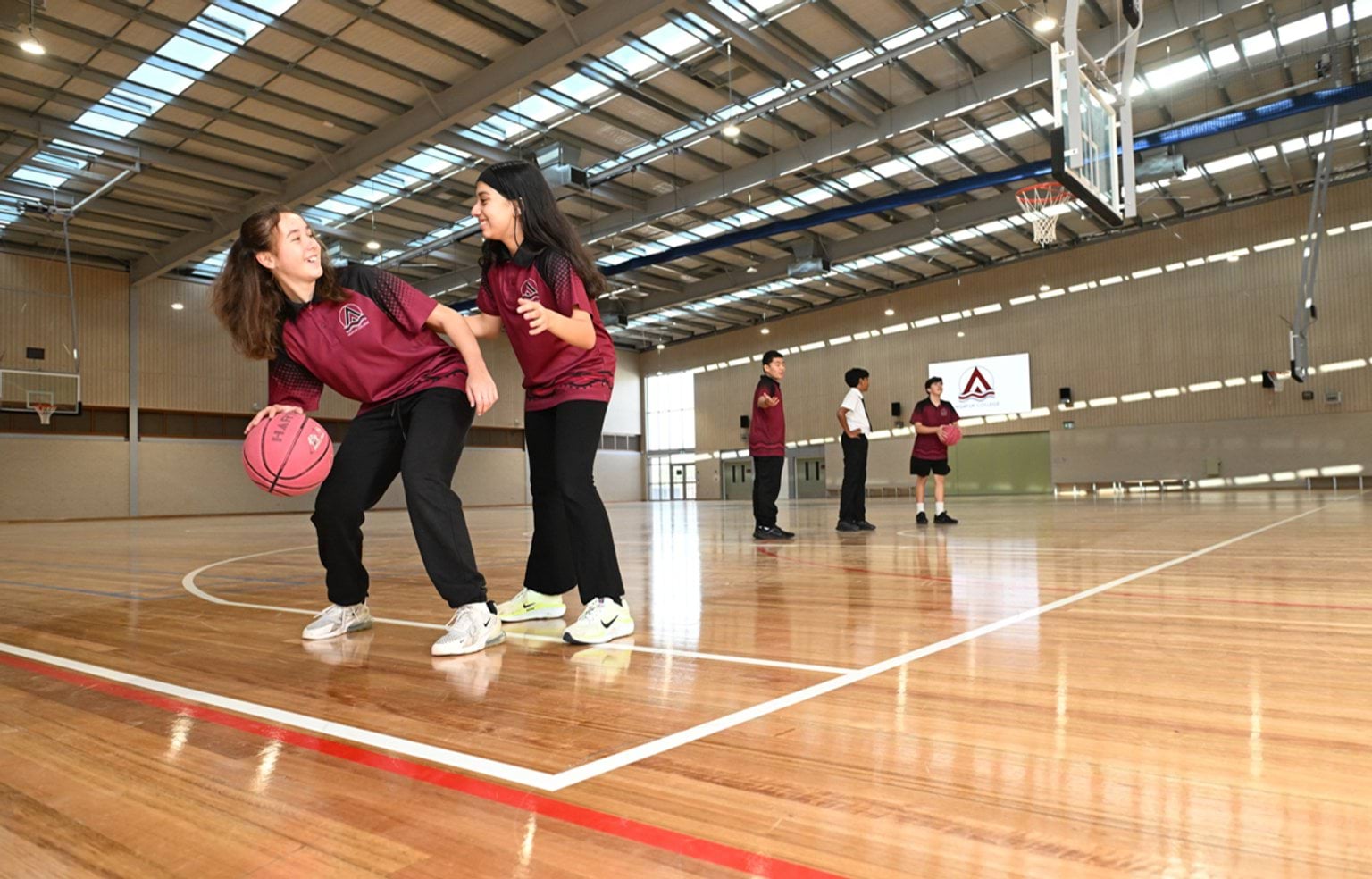 5 kids in maroon and black school uniform are playing with basketballs on an indoor covered basketball court
