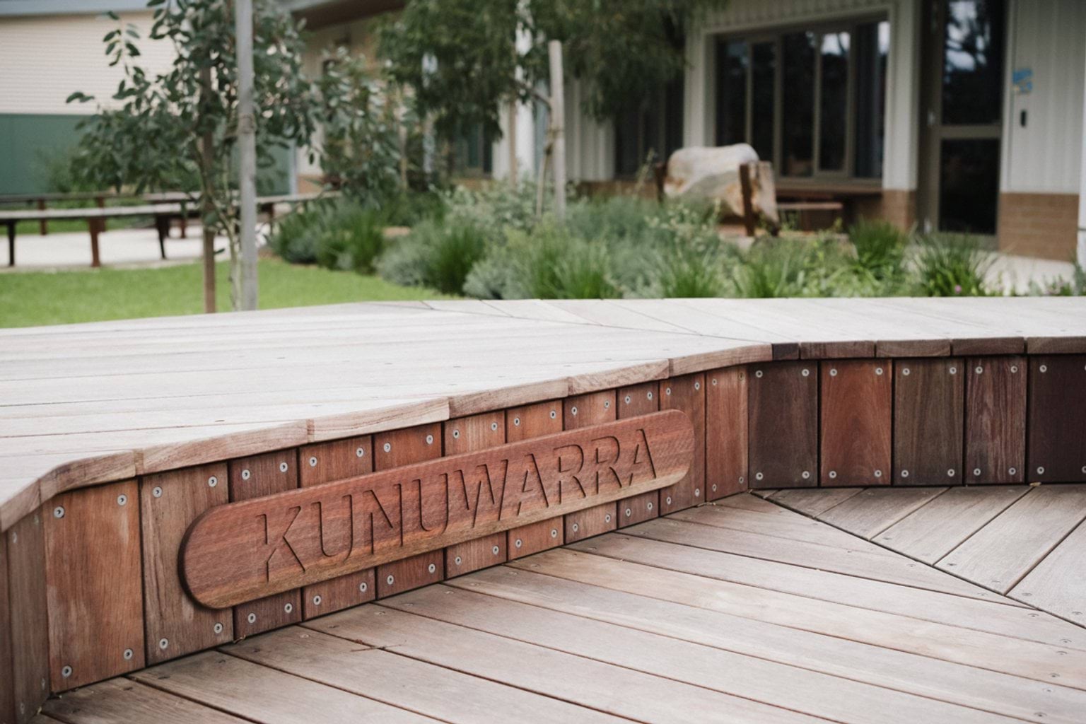 Outdoor wooden decked area with word 'Kunuwarra' engraved in wood on the side. We can see grass and garden area and a school building in the background