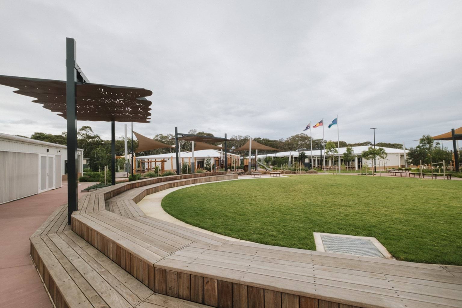 Outdoor grass oval with curved wooden benchtops surrounding. We can see a metal shelter, buildings and flagpoles in the background