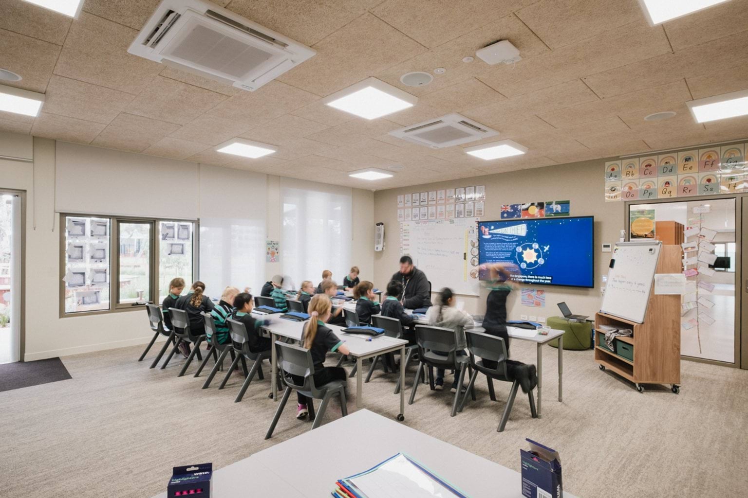 Students seated at desks in a classroom with cream carpet, a screen and whiteboard.