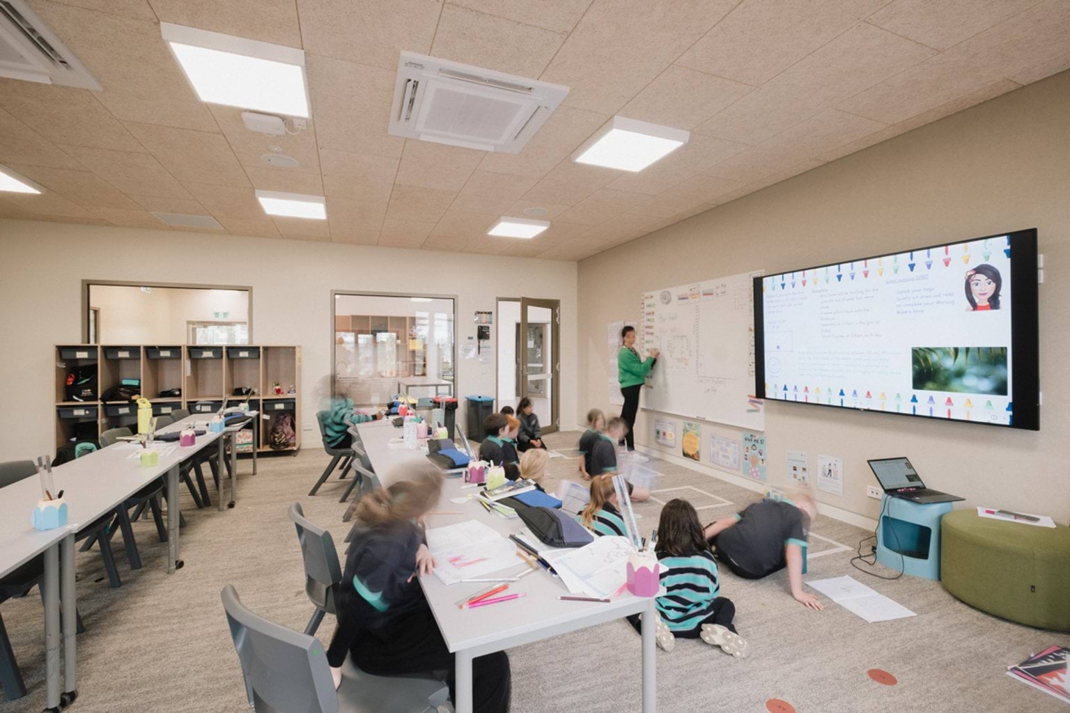 Students seated on chairs and on the floor while a teacher stands at the whiteboard. We can see desks, a screen and lockers along the far wall