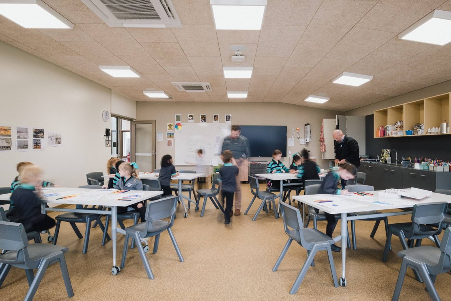 View from rear of classroom with students seated around tables and two teachers