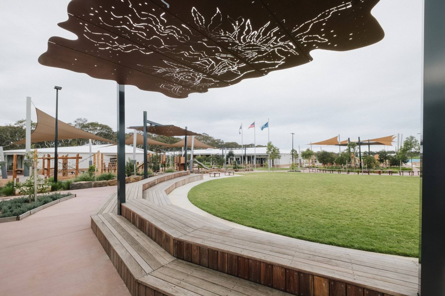 Oval grass area surrounded by curved wooden benches, a paved area and sheltered playground. We can see engraved artwork on a metal shelter above the benches