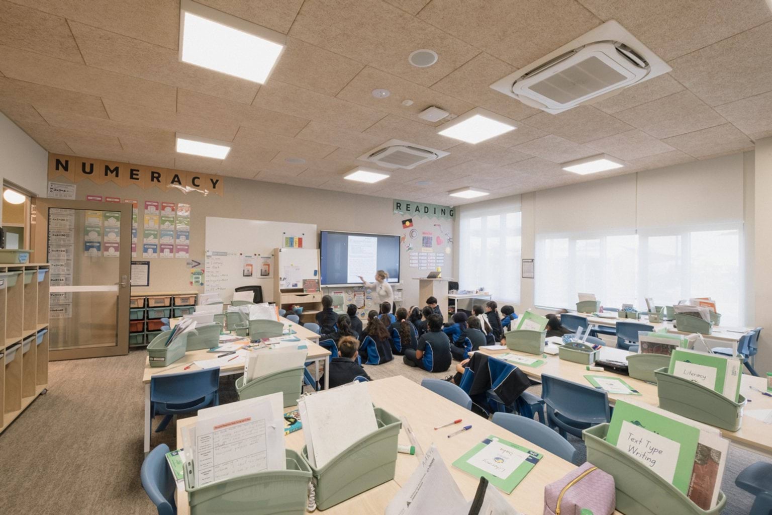 Classroom with chairs and tables with books and boxes. We can see children seated on the floor at the front of the classroom with the teacher in front of the screen