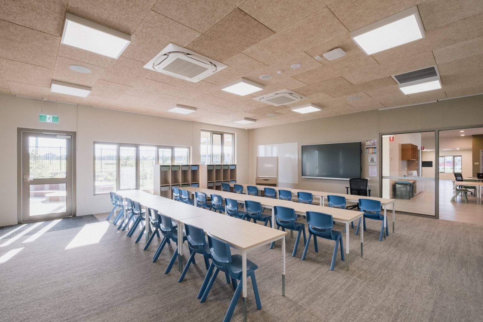 Classroom with grey carpet and rows of tables and chairs. We can see two doorways and windows along two walls as well as cubbies, a whiteboard and a screen