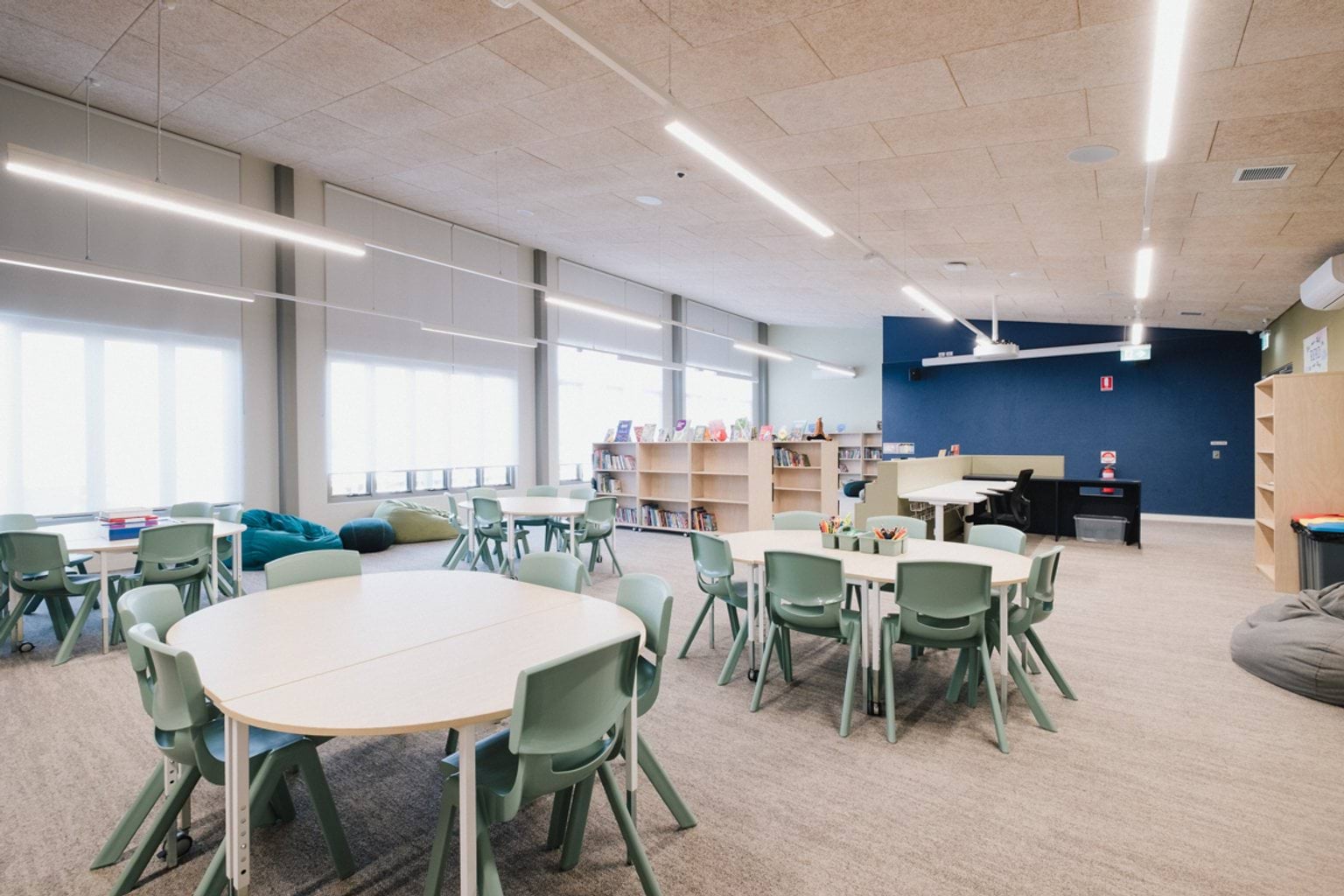 Classroom with grey carpet, tables and chairs throughout and a bookshelf and blue feature wall at the rear