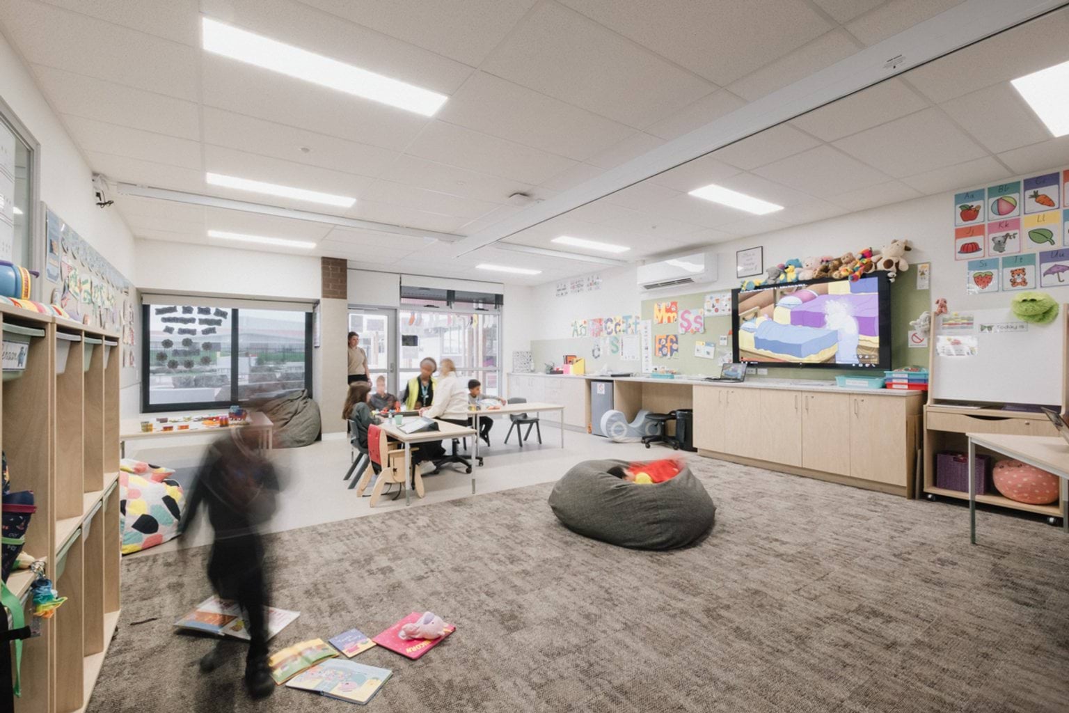 Classroom with grey patterned carpet, a screen and whiteboard with cabinets below and tables and chairs, lockers and a beanbag in the centre