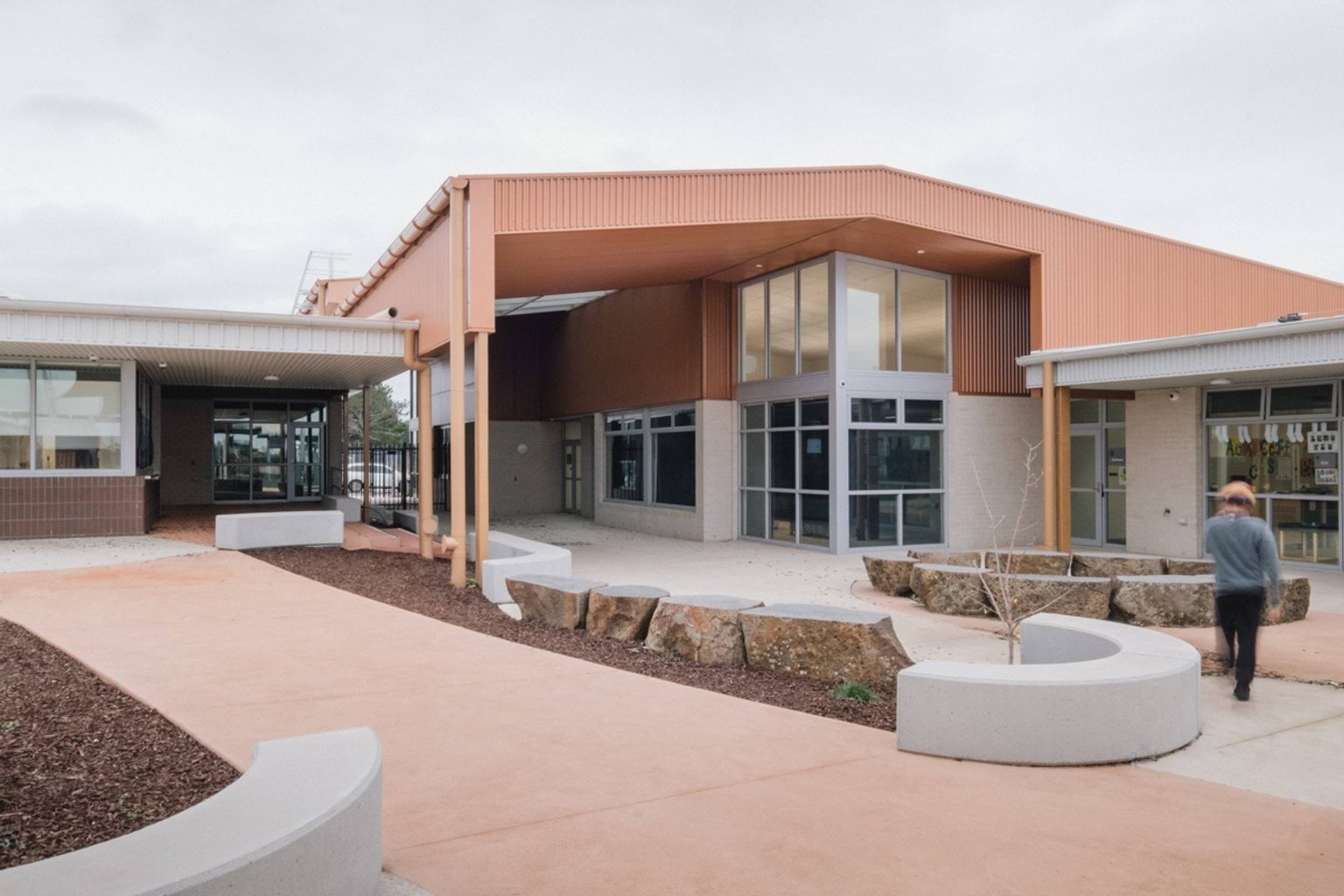 Paved entrance to two-storey orange and white building with garden beds, stone feature pieces and curved benches