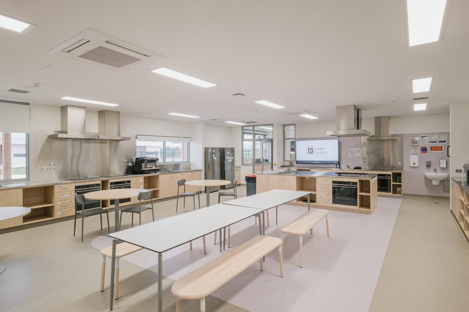 Food tech classroom with counter tops each fitted with extraction fans, sinks, ovens, stoves and microwaves. In the centre we can see a long bench with seats and rounded tables and chairs.
