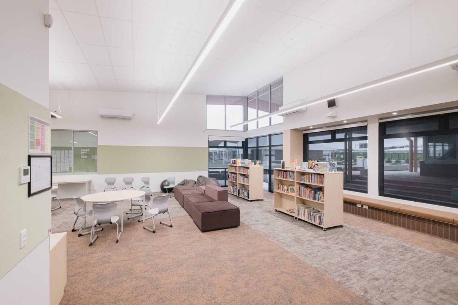 Classroom with grey carpet and light brown carpet and chairs at small round tables. We can see windows along the rear wall as well as a lounge seat and free-standing bookshelves