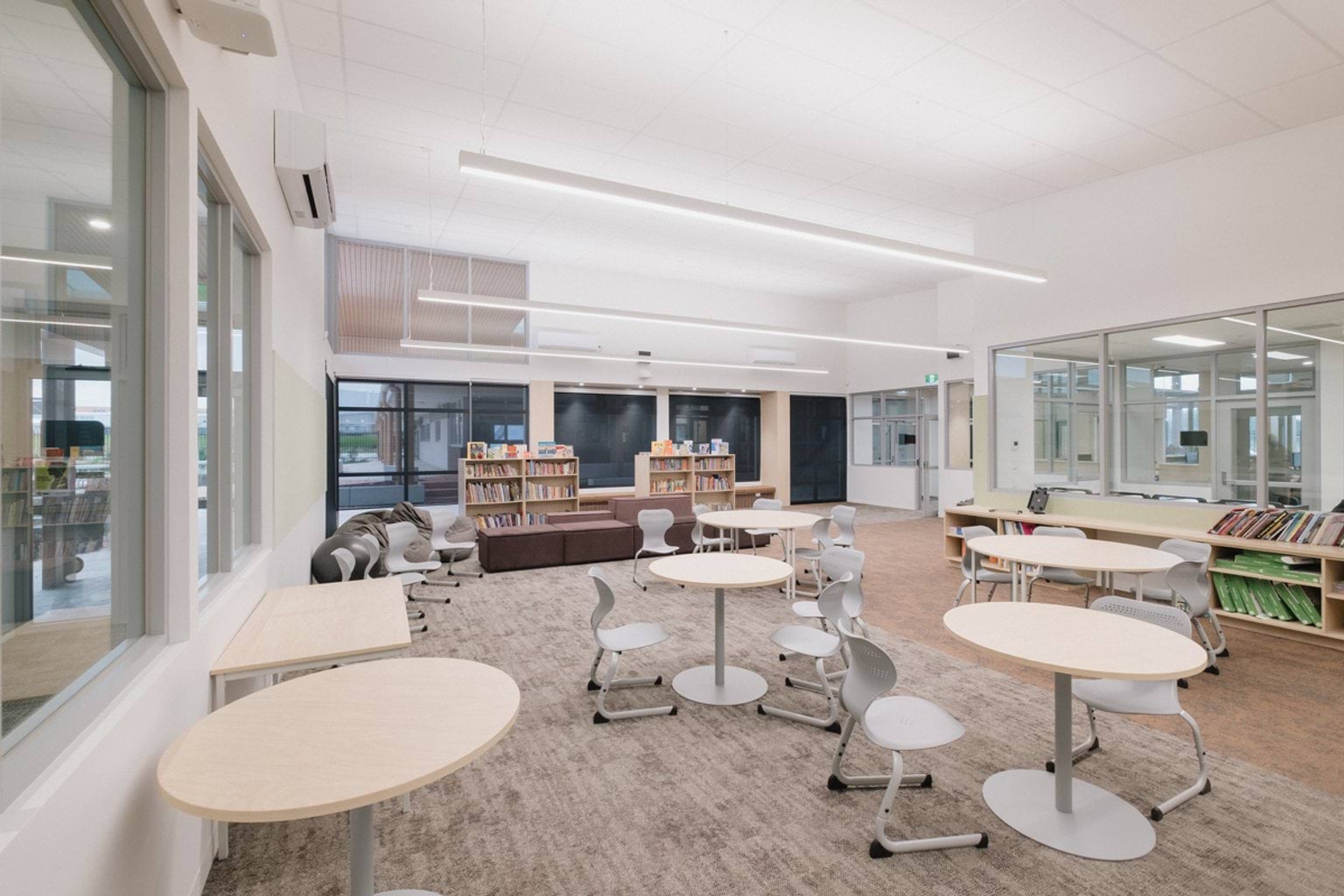 Classroom with grey carpet and light brown carpet and chairs at small round tables. We can see windows and bookshelves along all walls and beanbags in the far corner.
