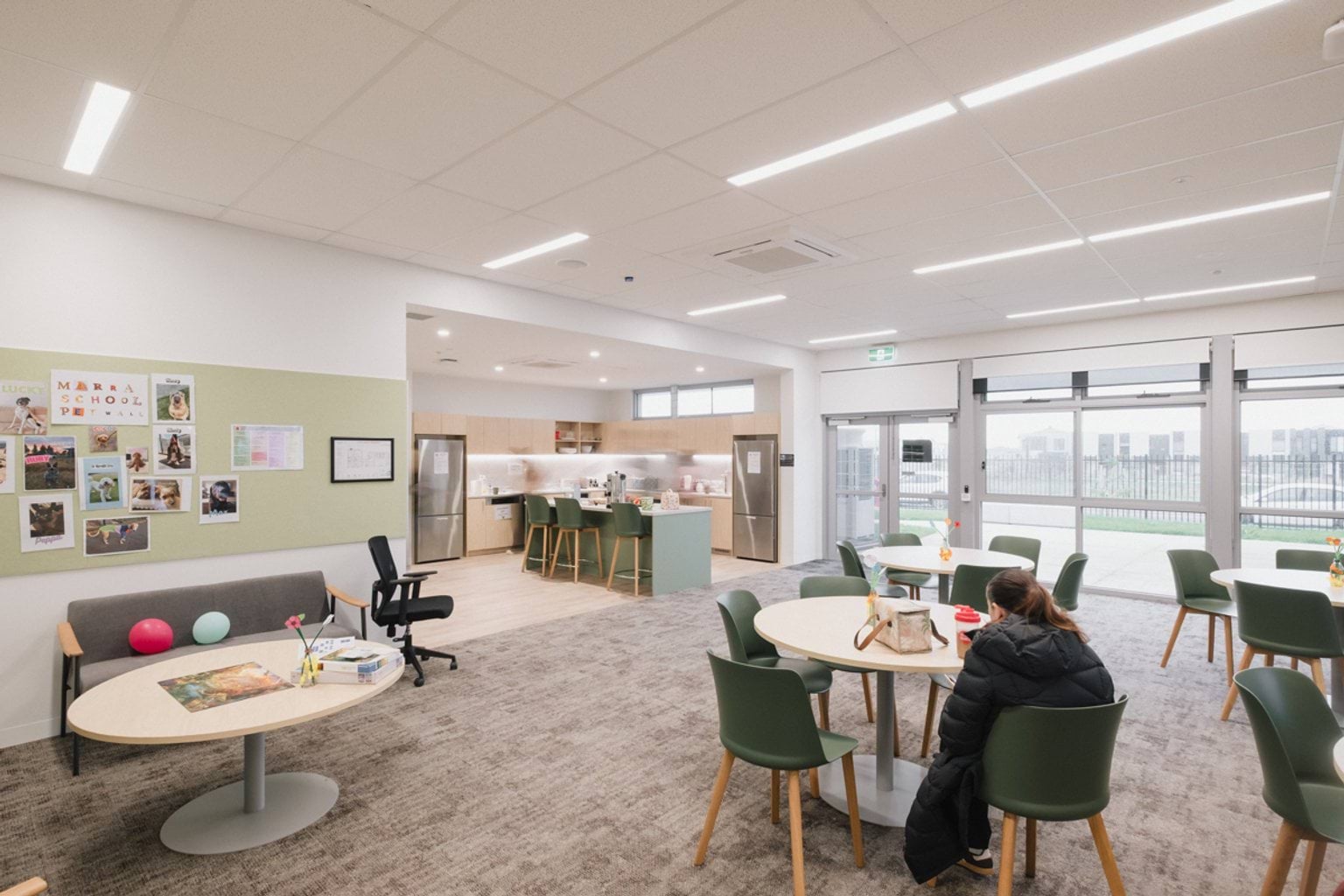 Open plan room with tables and chairs in carpeted area, a couch and round coffee table. In the rear of the room we can see an attached kitchen space