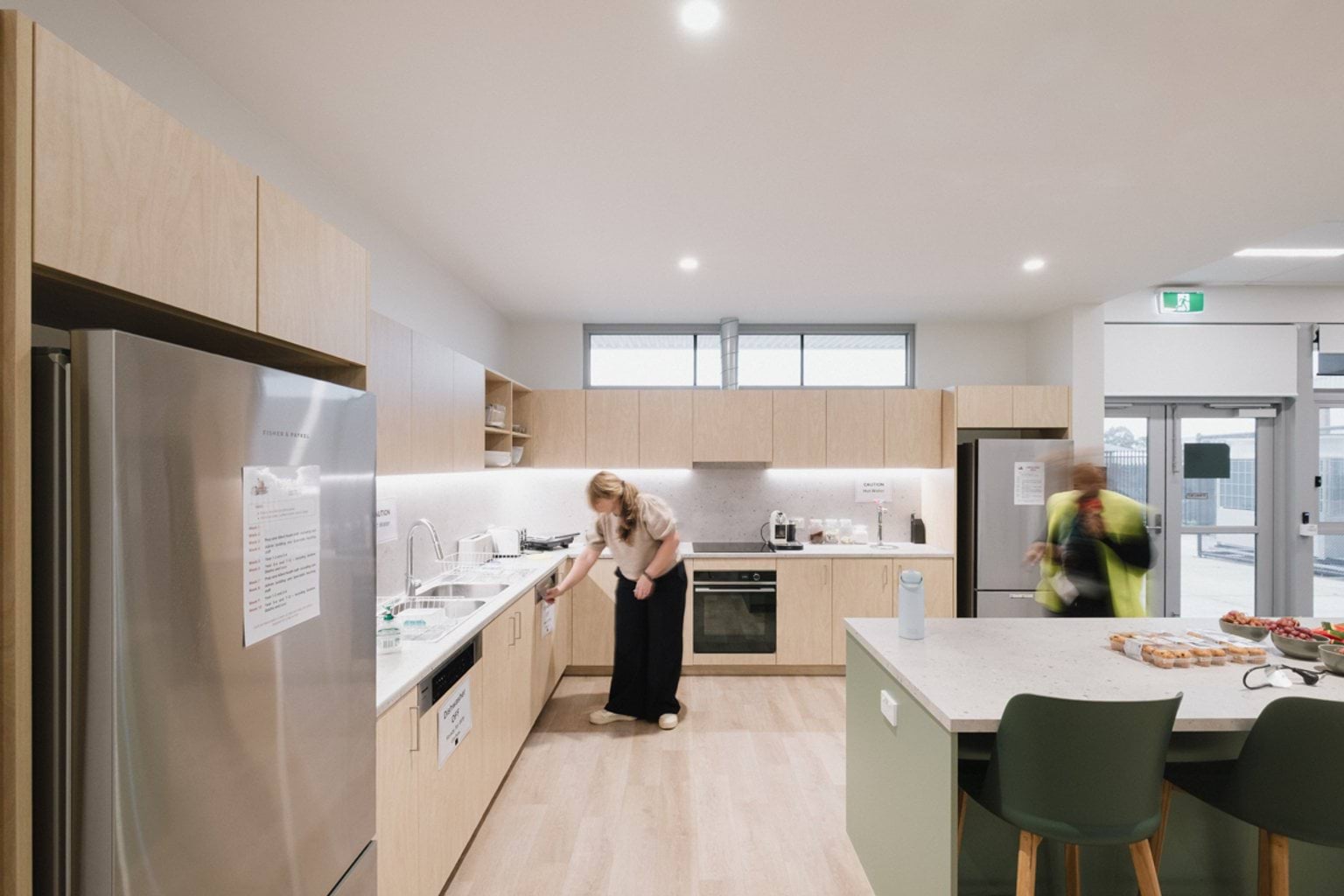 Open plan kitchen space with island benchtop, green chairs, fridges, sink and stove. We can see two adults making use of the kitchen.