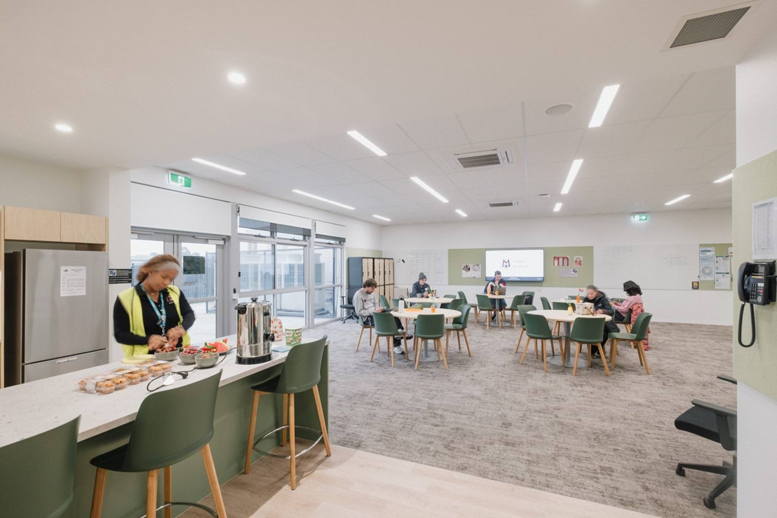 Large room with kitchen benchtop, chairs, fridge and carpeted leisure area with tables and chairs. We can see people seated around the tables and a person standing at the kitchen benchtop.