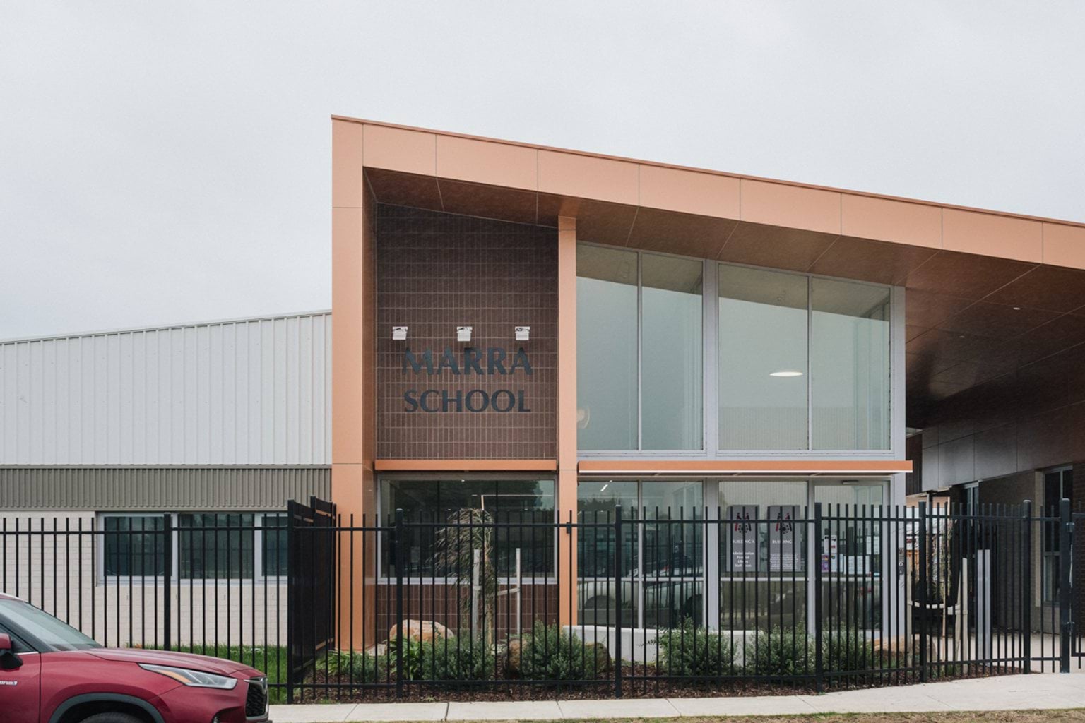 Gated building entrance with orange beams covering two-storey building. We can see floor to ceiling windows and the letters 'Marra School' in black on the building wall
