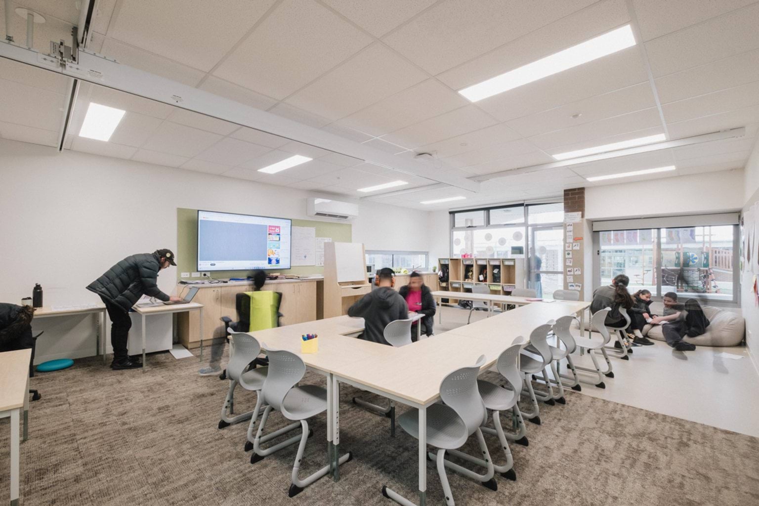 Classroom with grey patterned carpet, a screen and whiteboard with tables and chairs throughout