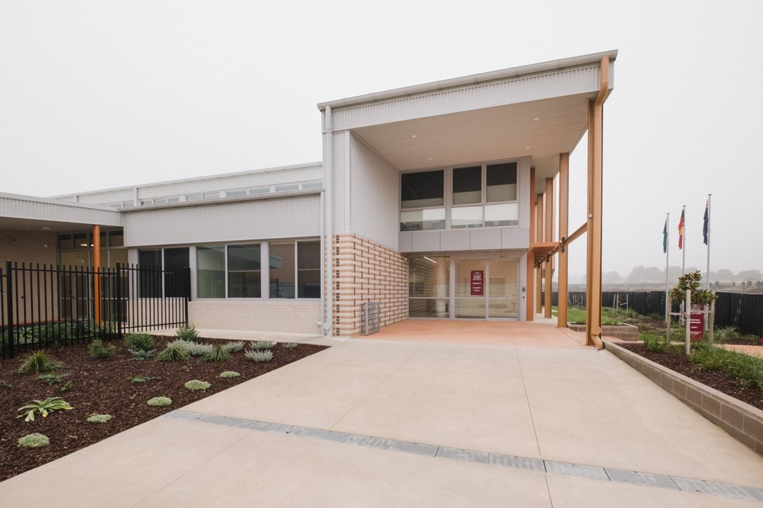 Paved entrance to two-storey building with garden beds on both sides. We can see three flag poles in the distance with grey cloud cover