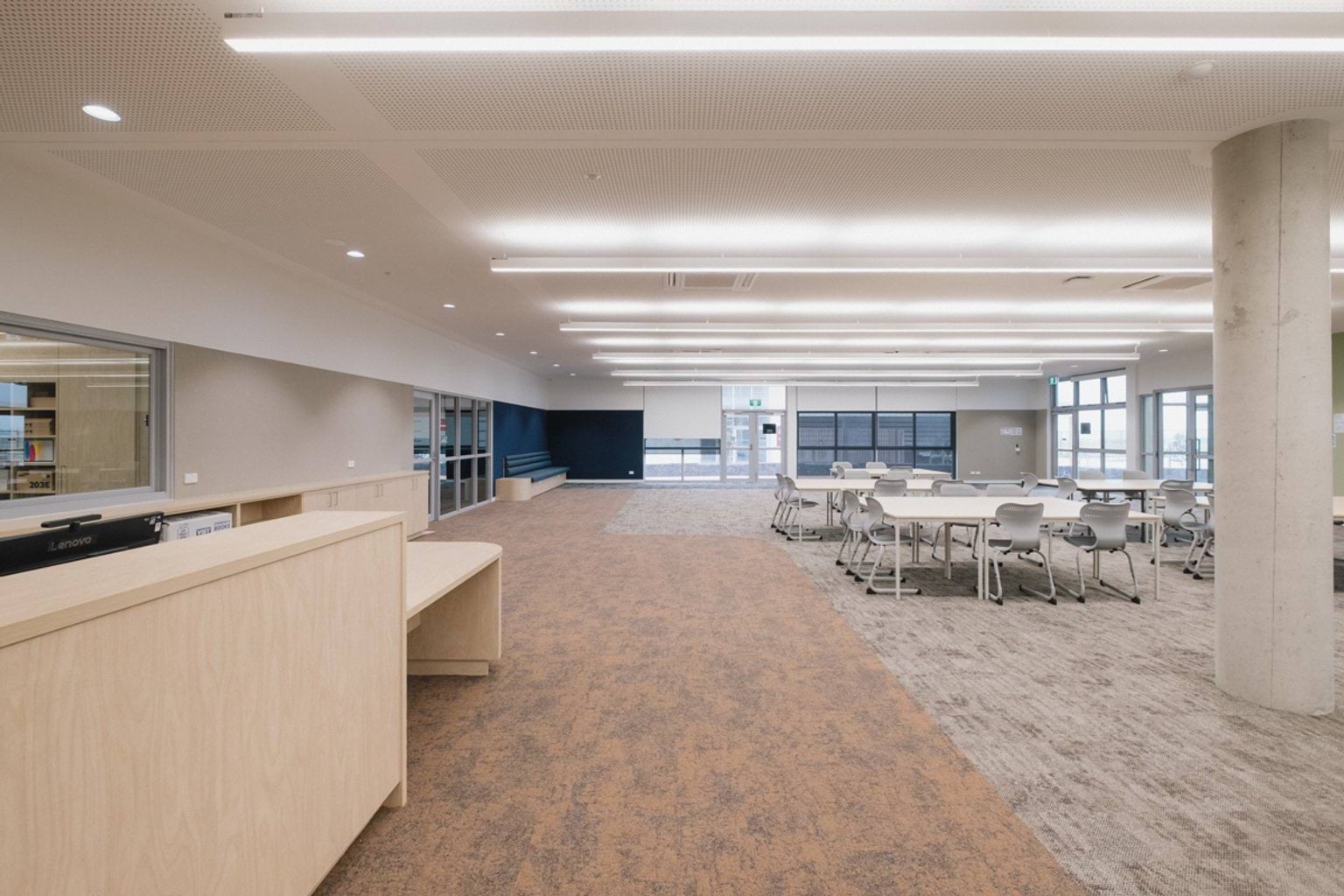 Open plan learning area with orange and grey carpet, tables and chairs in the centre and a desk on the left hand side. We can see windows at the rear of the room and windows into surrounding rooms